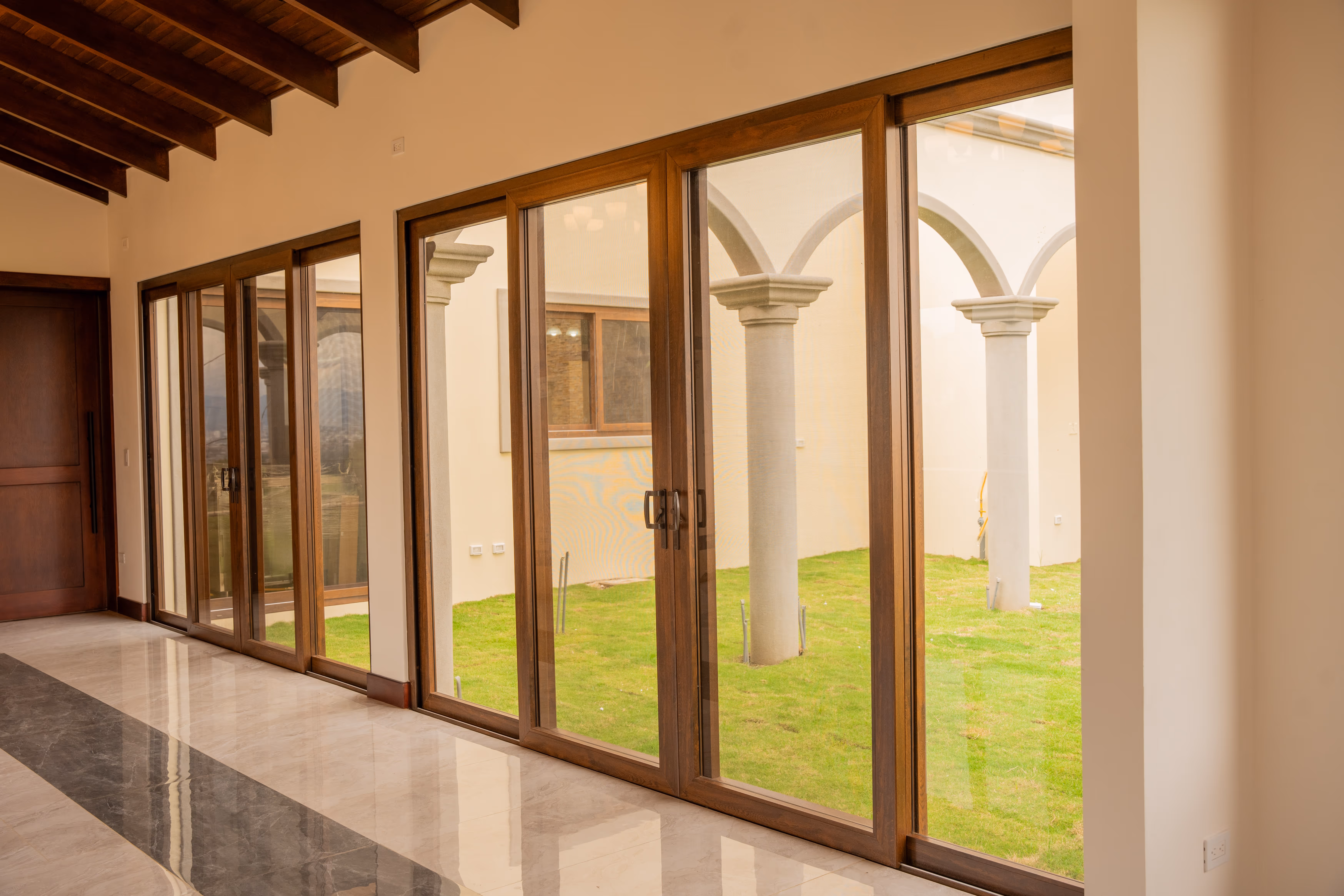 Interior de una habitación con suelos de mármol y puertas corredizas de cristal con marco de PVC imitación madera que dan a un patio con columnas y césped verde.