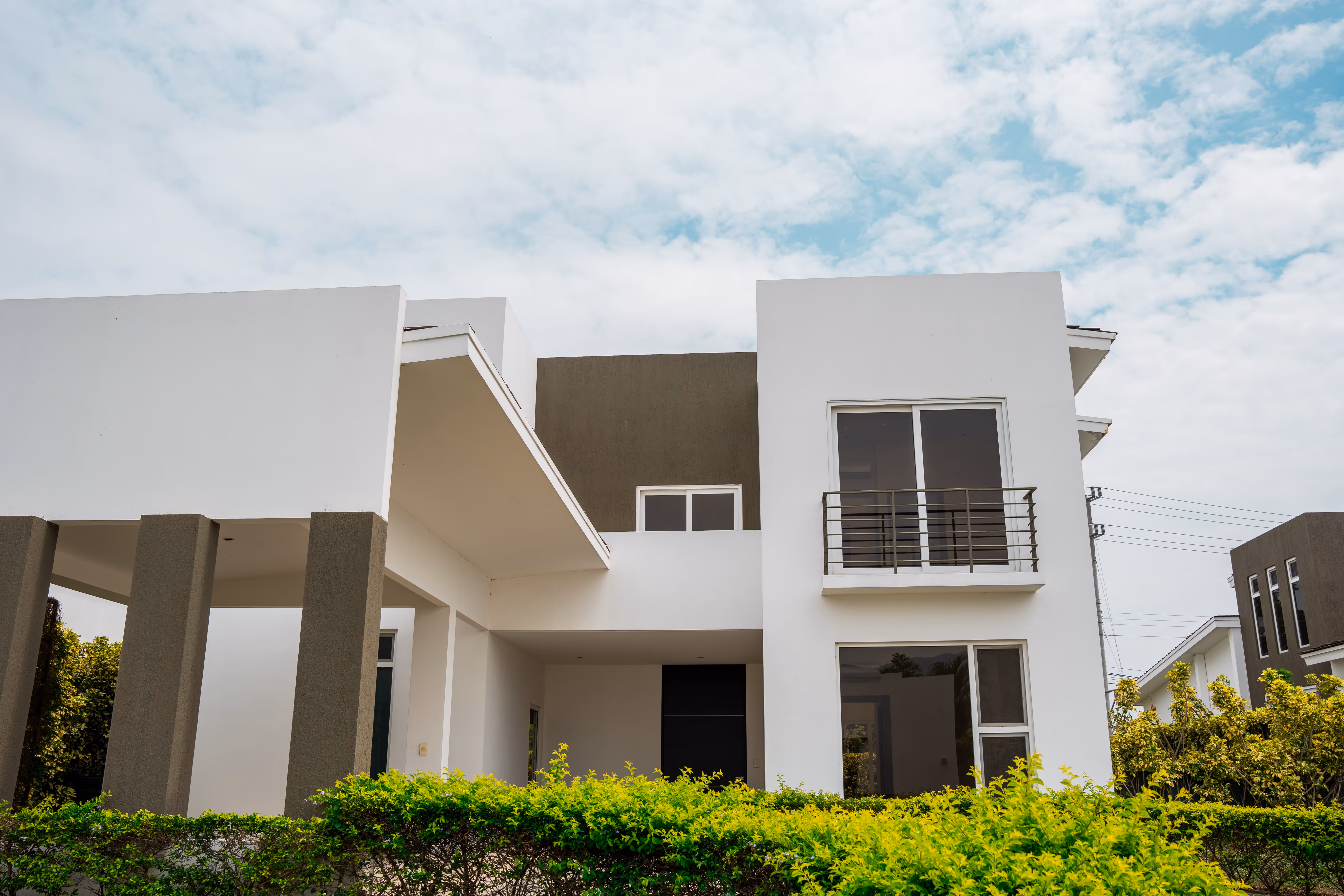 Casa moderna de dos pisos con fachada blanca y gris, con puertas y ventanas de vidrio con marcos de pvc blanco. con balcones y arbustos verdes al frente bajo un cielo parcialmente nublado.