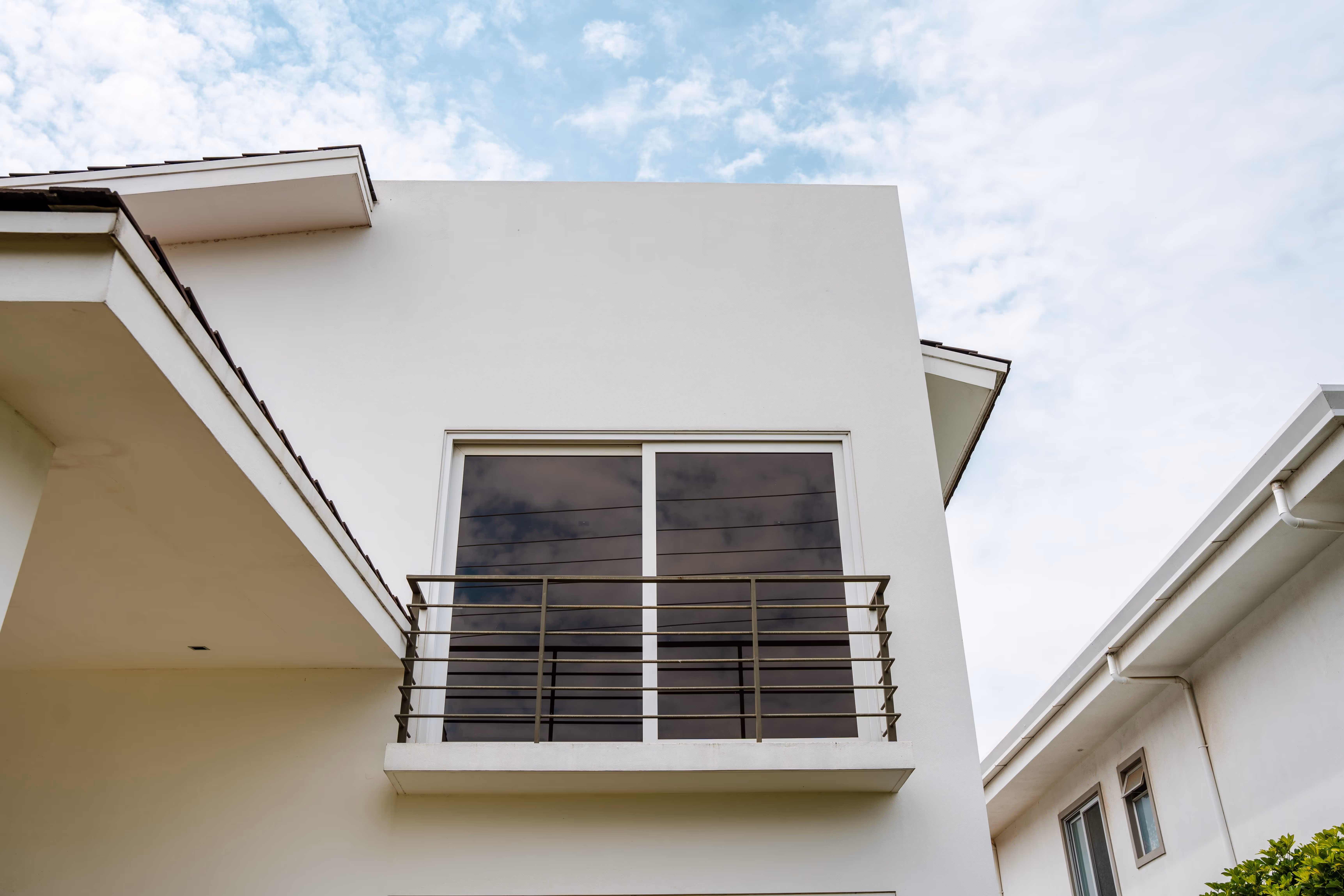 Balcón pequeño con barandilla metálica en frente de puerta corrediza de vidrio con marcos de pvc blanco. en una fachada blanca de casa moderna bajo cielo parcialmente nublado.