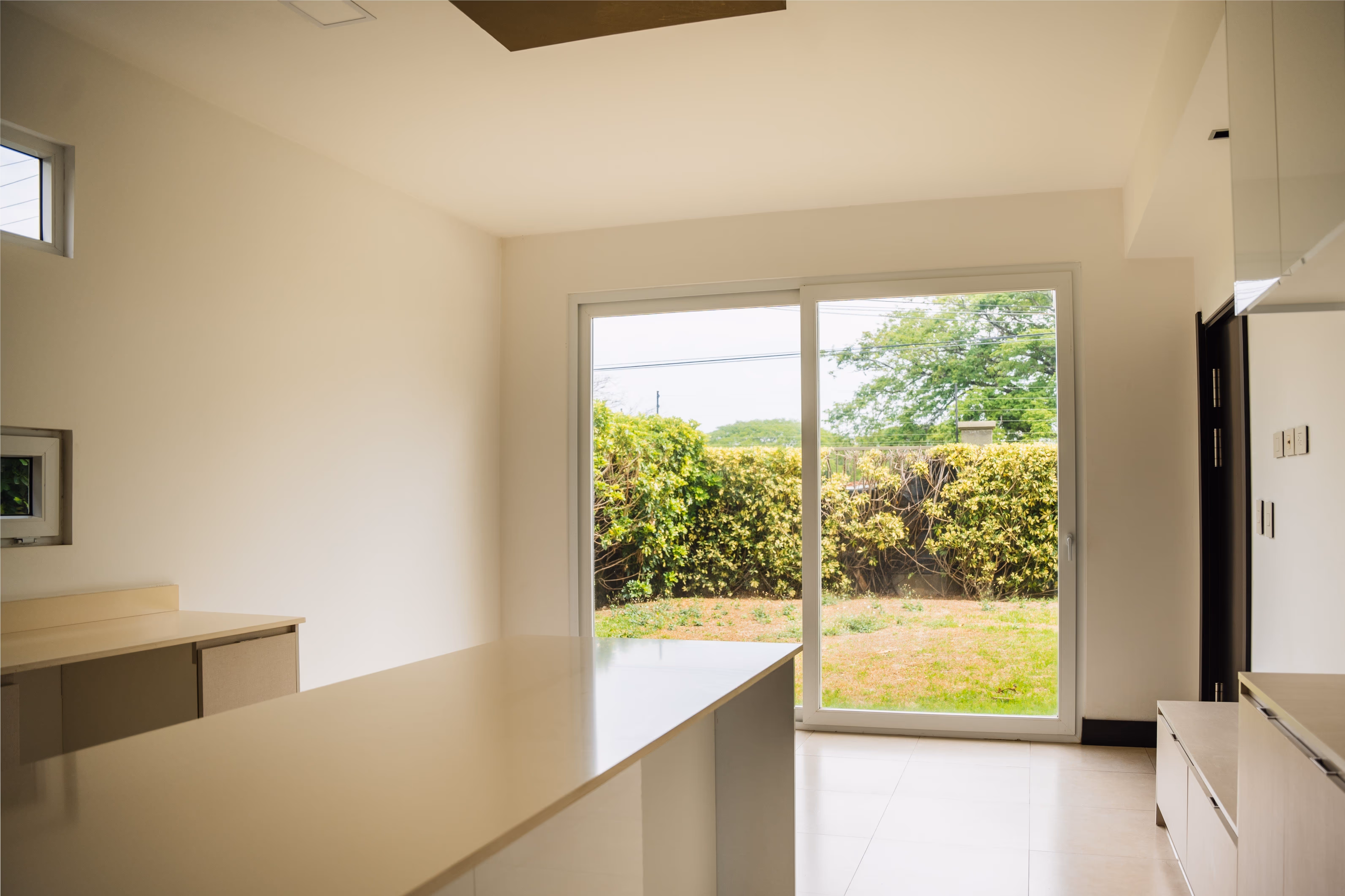 Interior luminoso de una cocina moderna con encimera blanca y puerta corrediza de vidrio con marcos de pvc blanco que muestra un jardín exterior con arbustos.