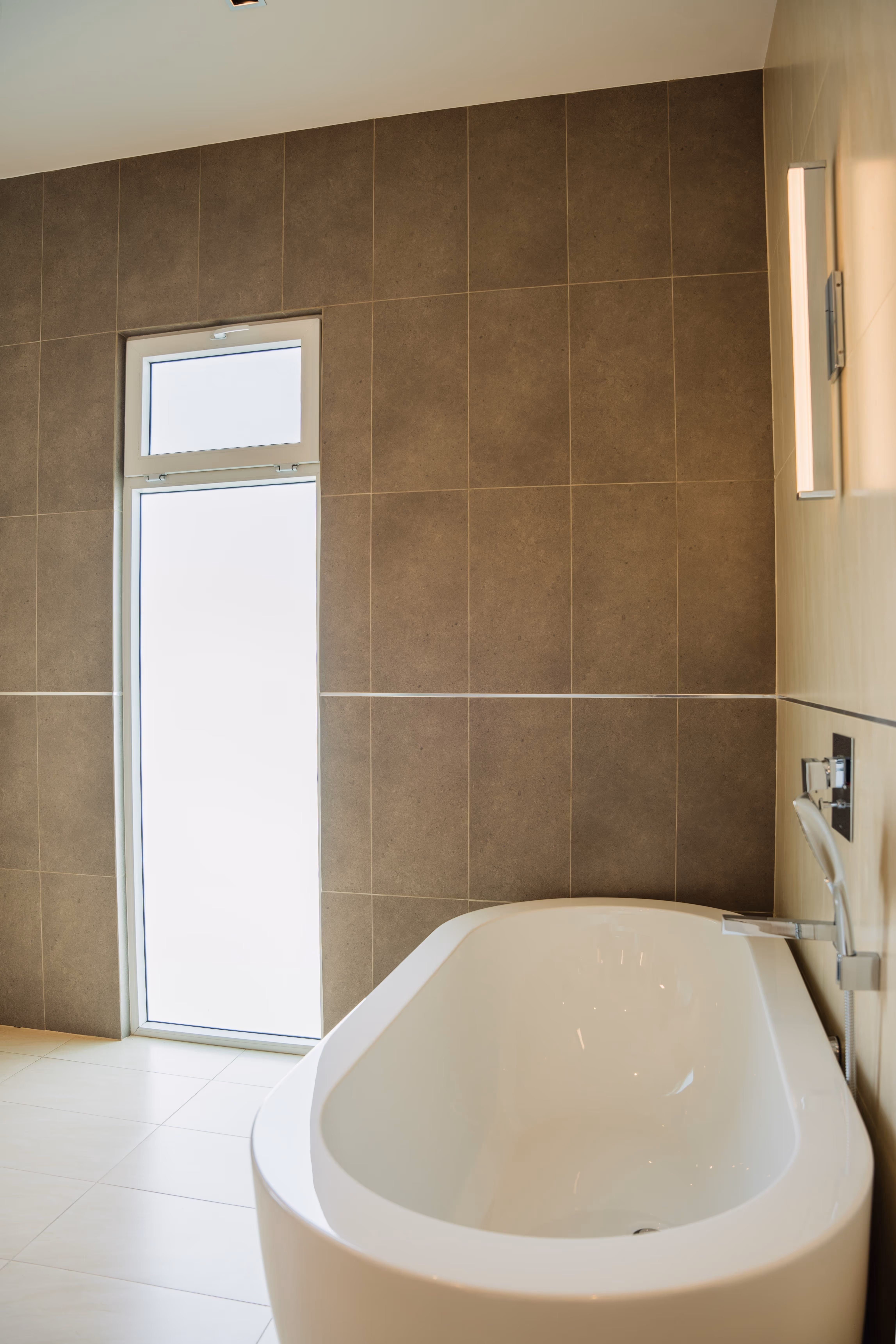 Baño moderno con bañera blanca independiente, pared de azulejos marrones y ventana alta rectangular en pvc blanco con vidrio arenado.