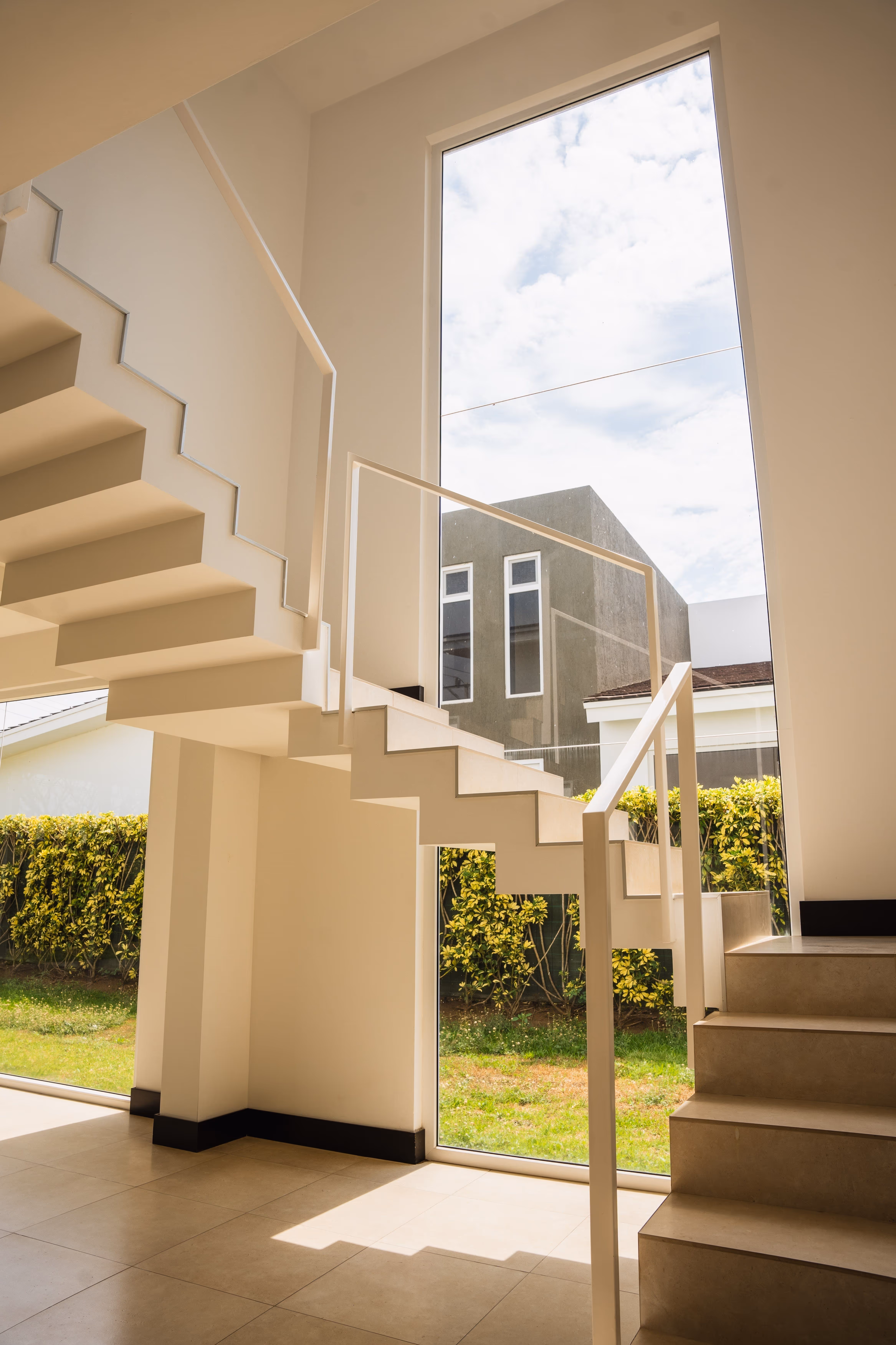 Escaleras modernas de interior con barandilla blanca junto a una gran ventana de pvc blanco que muestra césped y arbustos afuera.