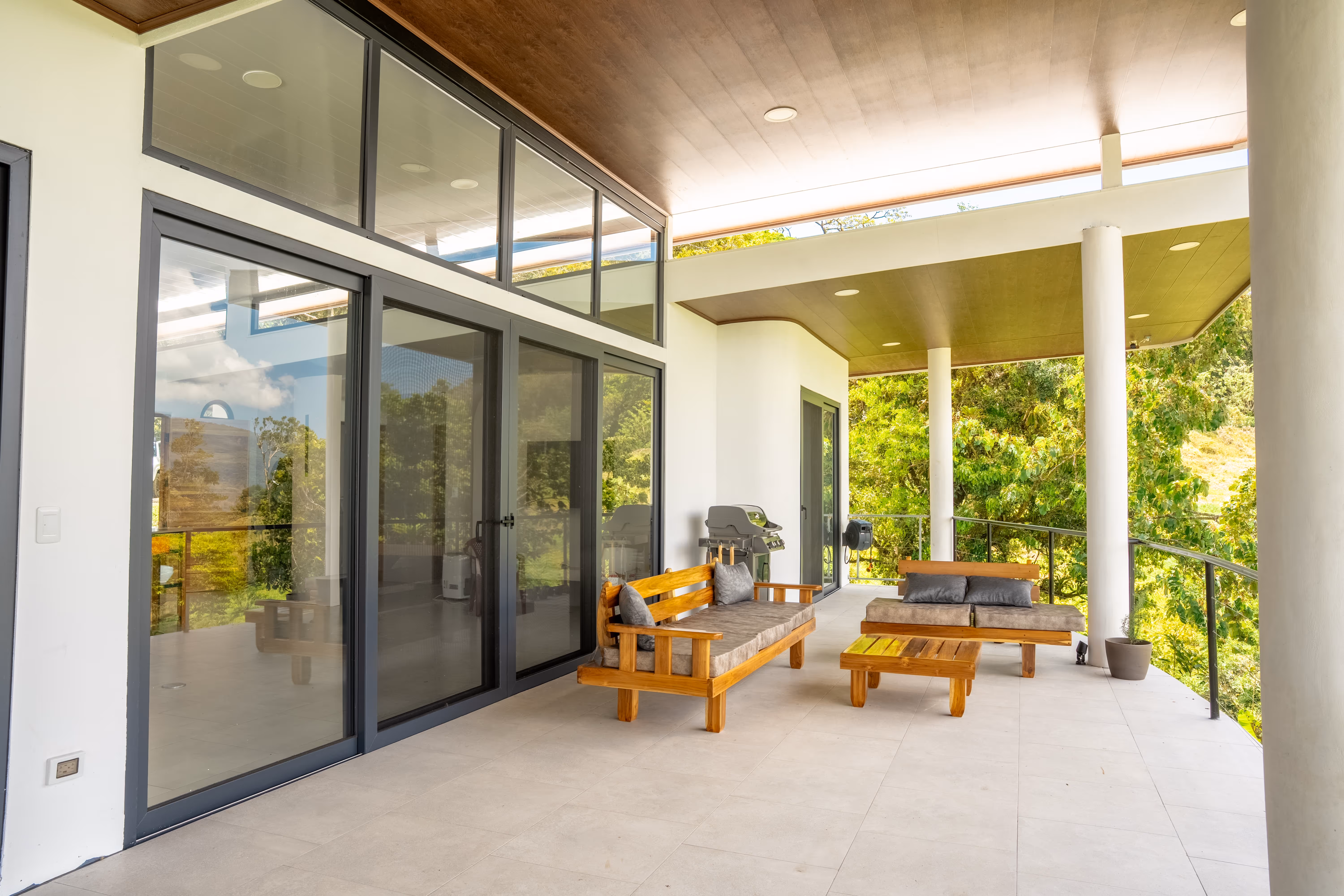 Terraza amplia con muebles de madera, puertas corredizas en pvc gris, cojines grises, parrilla y barandilla con vista a vegetación verde.