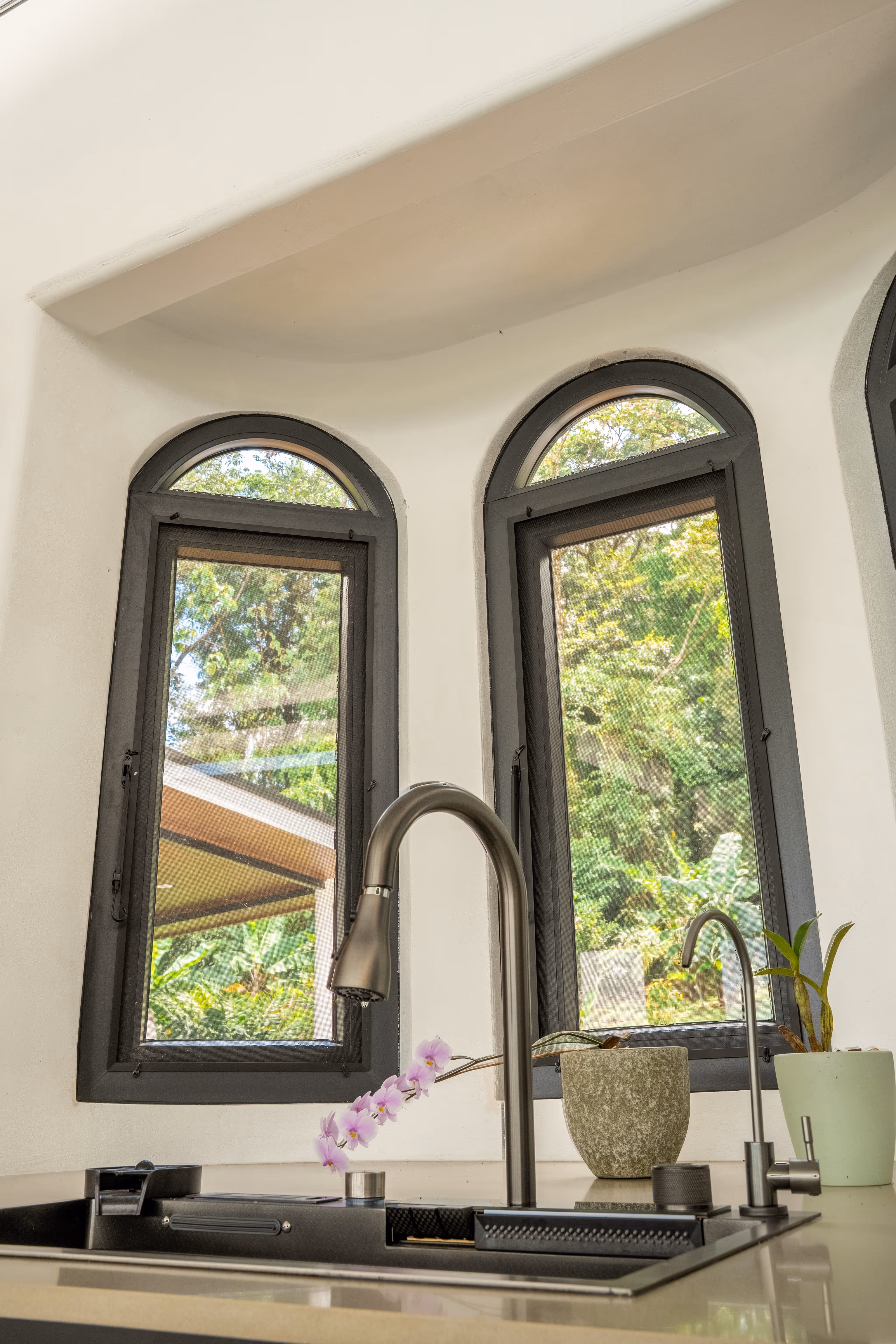 Fregadero moderno con grifería de acero inoxidable frente a tres ventanas arqueadas de pvc gris con vista a árboles y plantas.