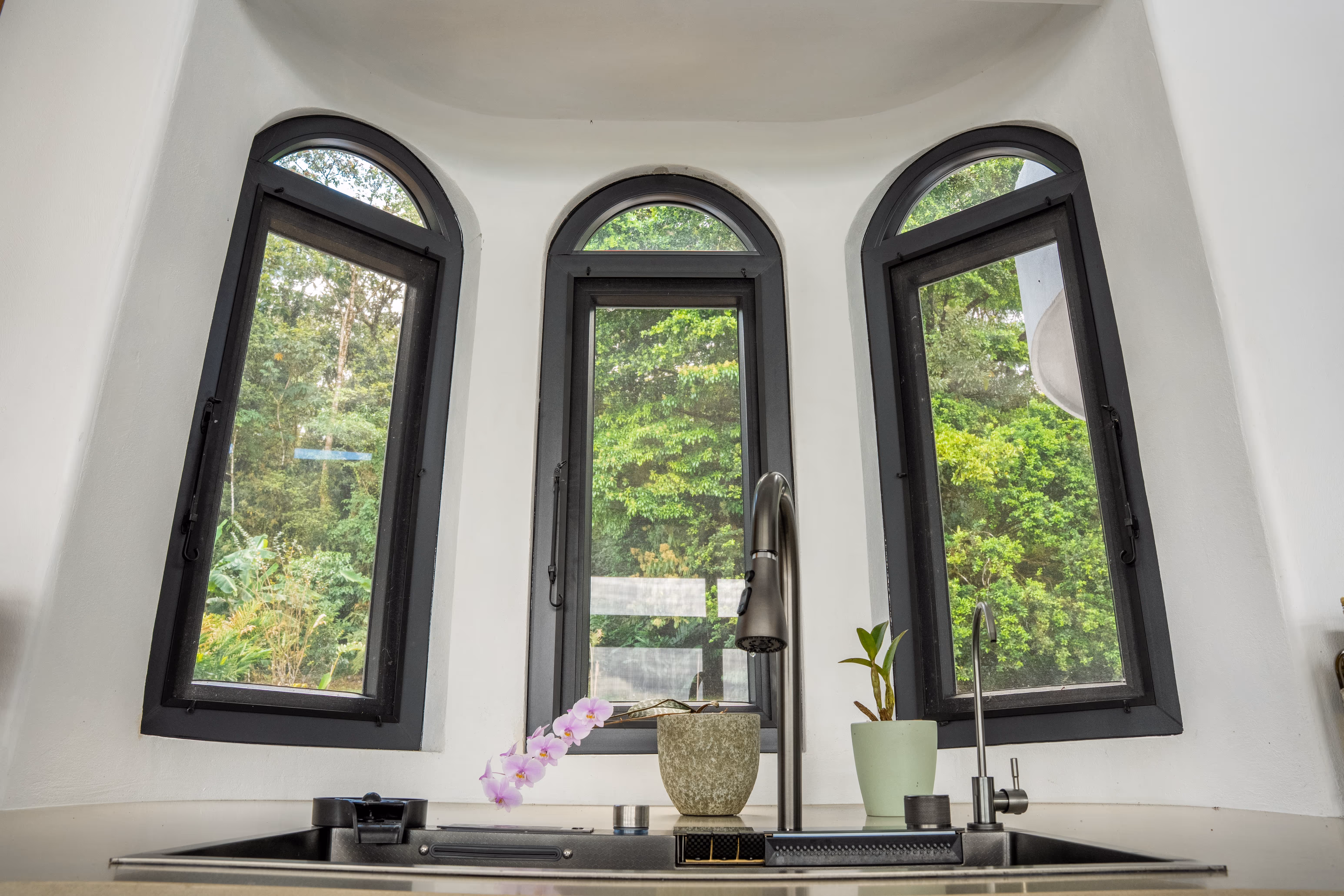 Ventanas arqueadas de pvc gris sobre fregadero con grifo y dos plantas en macetas, con vista a árboles verdes.
