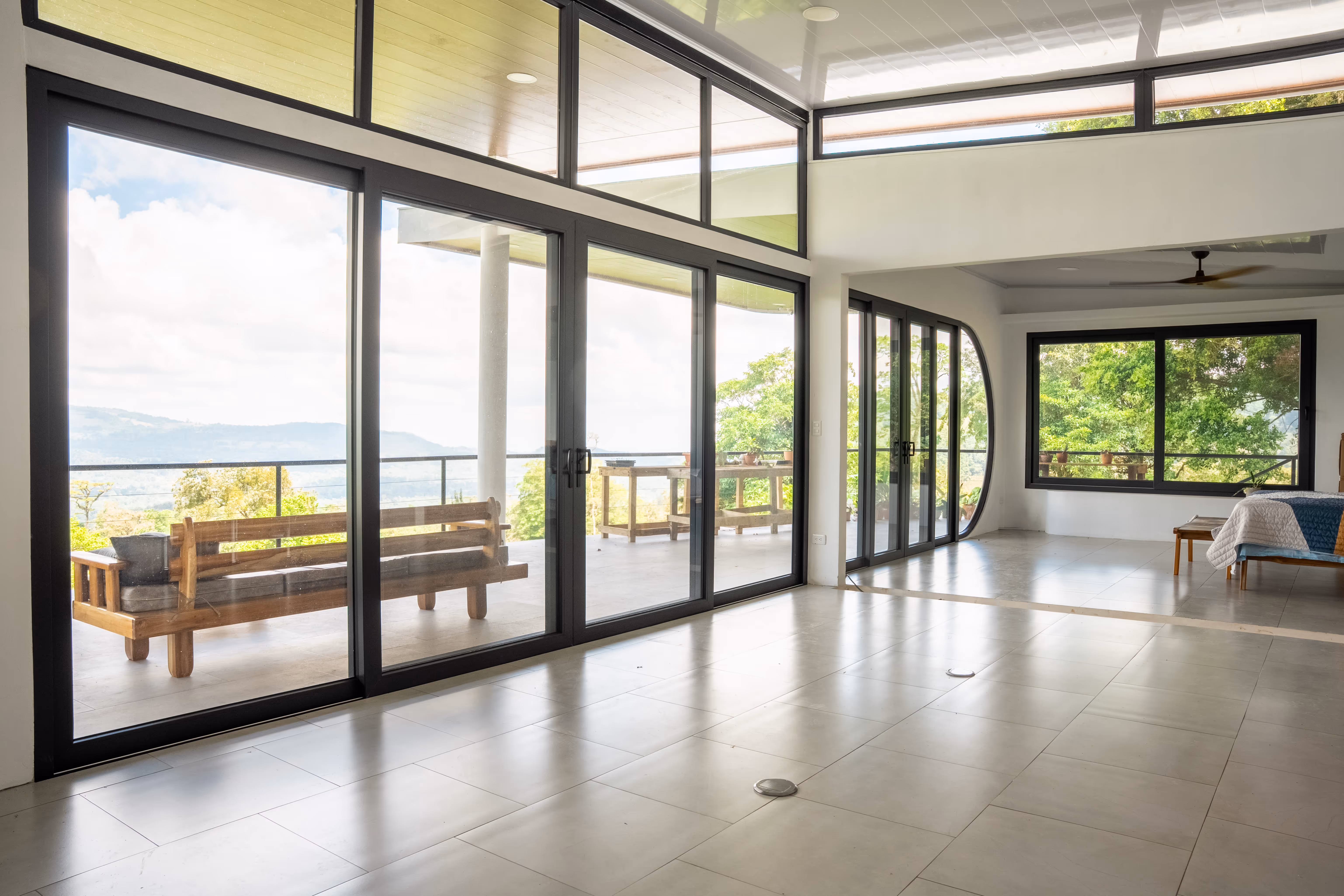 Amplia sala con grandes ventanales de vidrio  en pvc gris y puerta corrediza que muestran un balcón con banco de madera y vistas a un paisaje montañoso y árboles verdes.