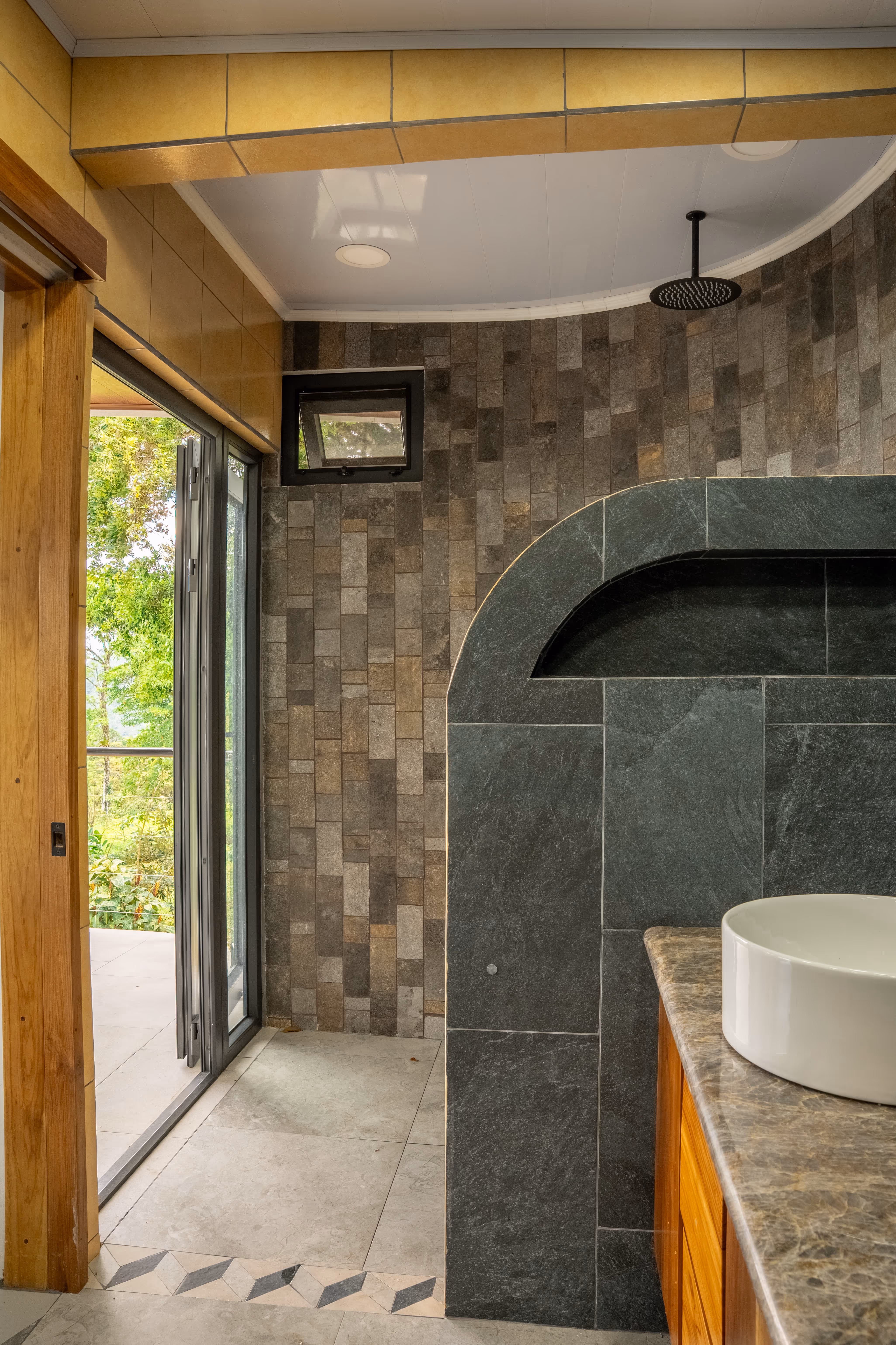 Baño moderno con ducha de lluvia, paredes de azulejos grises, fregadero blanco sobre encimera de mármol y puerta de pvc gris abierta con vista al jardín.