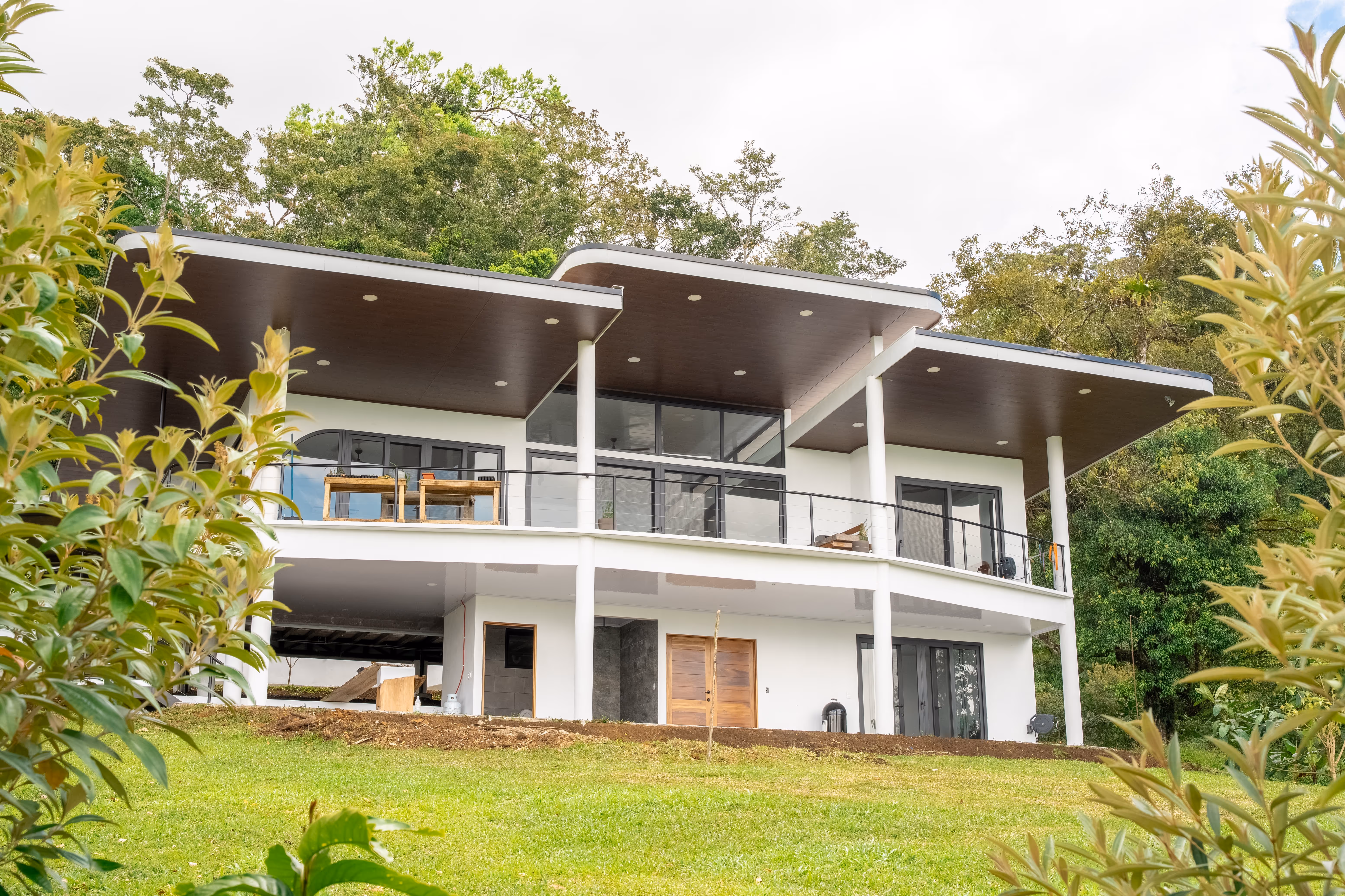 Casa moderna de dos pisos con fachada blanca, puertas y ventanas de pvc gris techo oscuro y balcones con barandales de metal, rodeada de césped verde y árboles.
