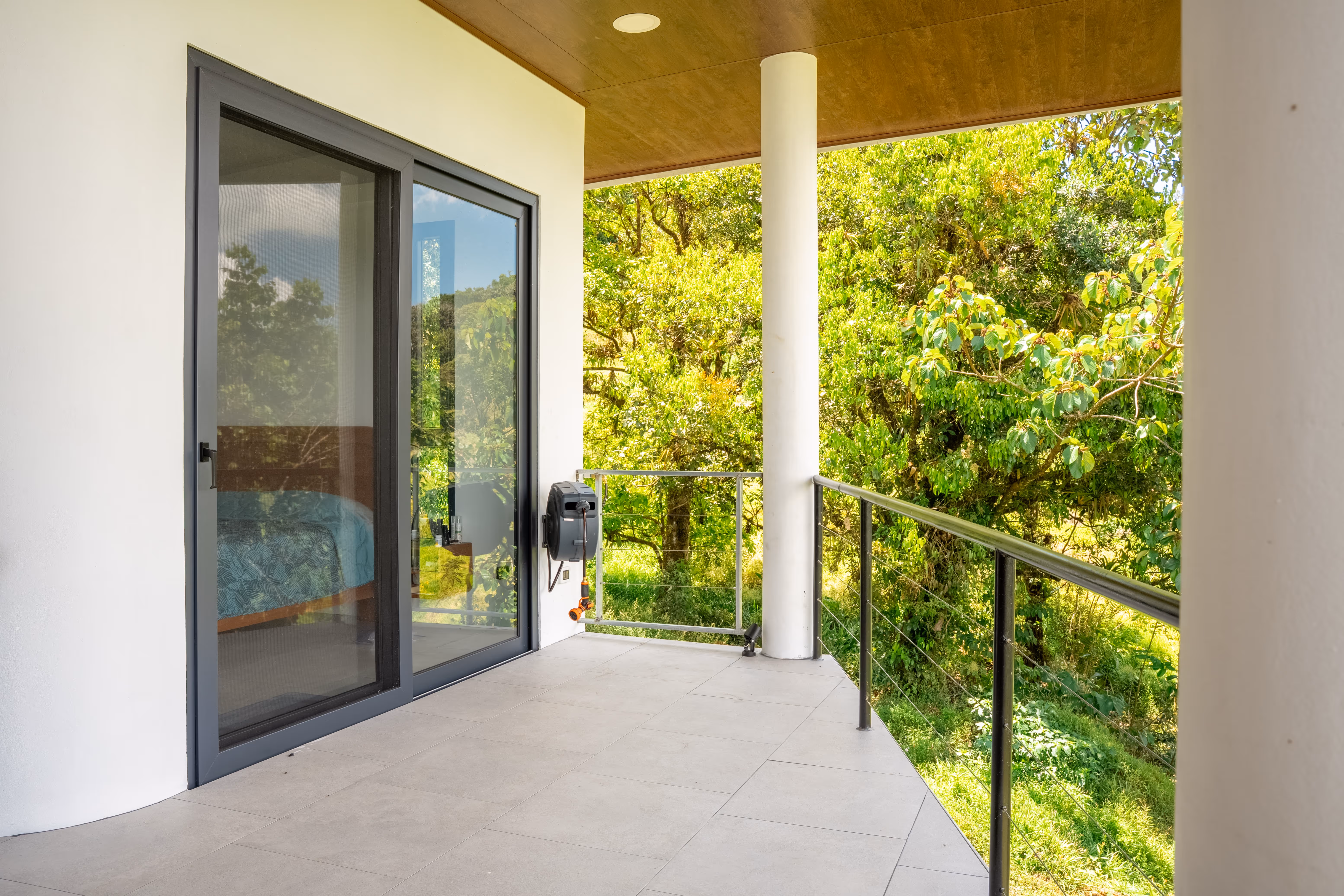 Balcón con piso de baldosas, barandilla metálica y puerta corrediza de pvc gris con vista a árboles verdes.