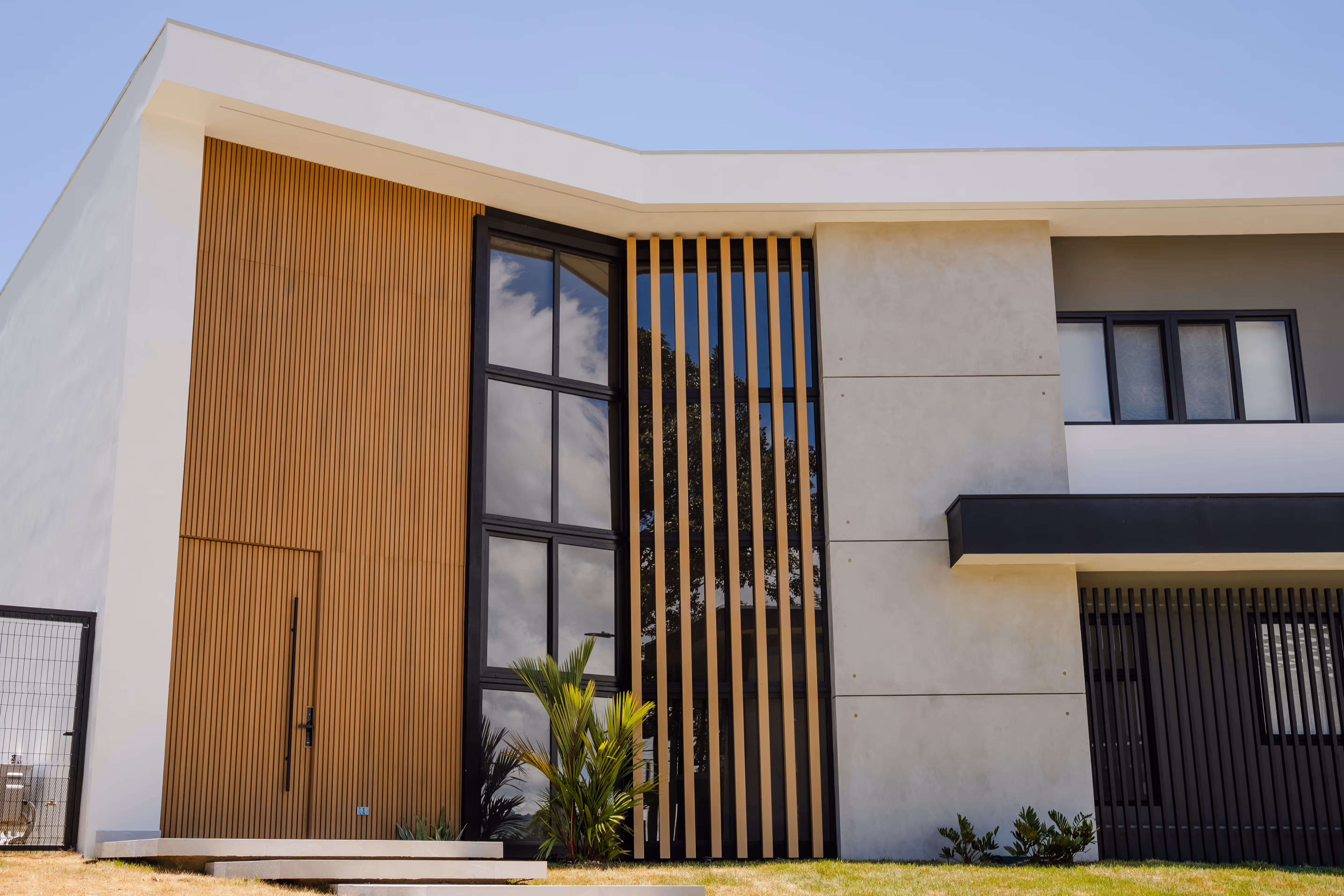 Fachada moderna de casa con paneles de madera, ventanas grandes de pvc negro y jardín con césped y plantas.