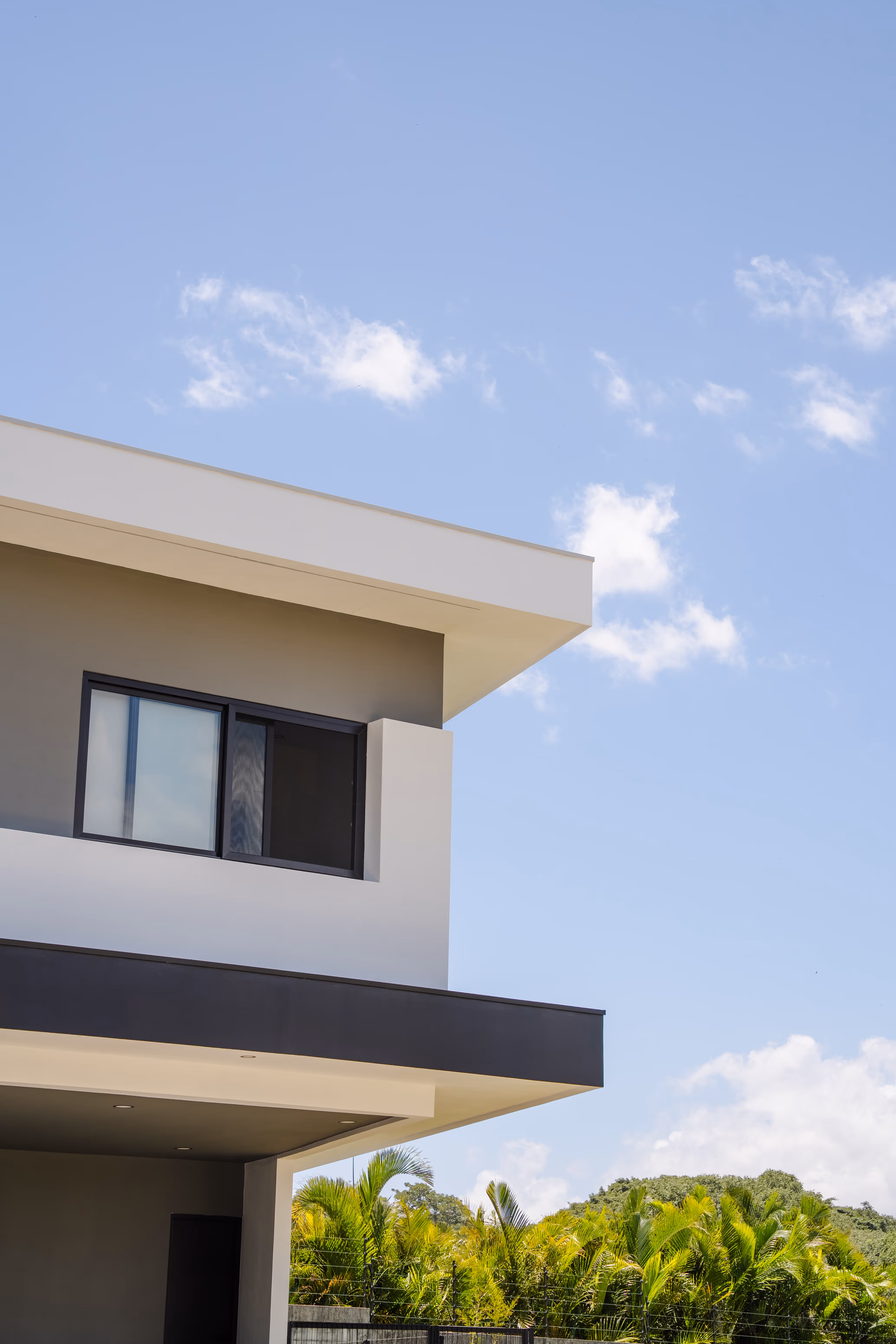 Esquina de una casa moderna de dos pisos con ventanas grandes corredizas de pvc negro, cielo azul y vegetación verde alrededor.
