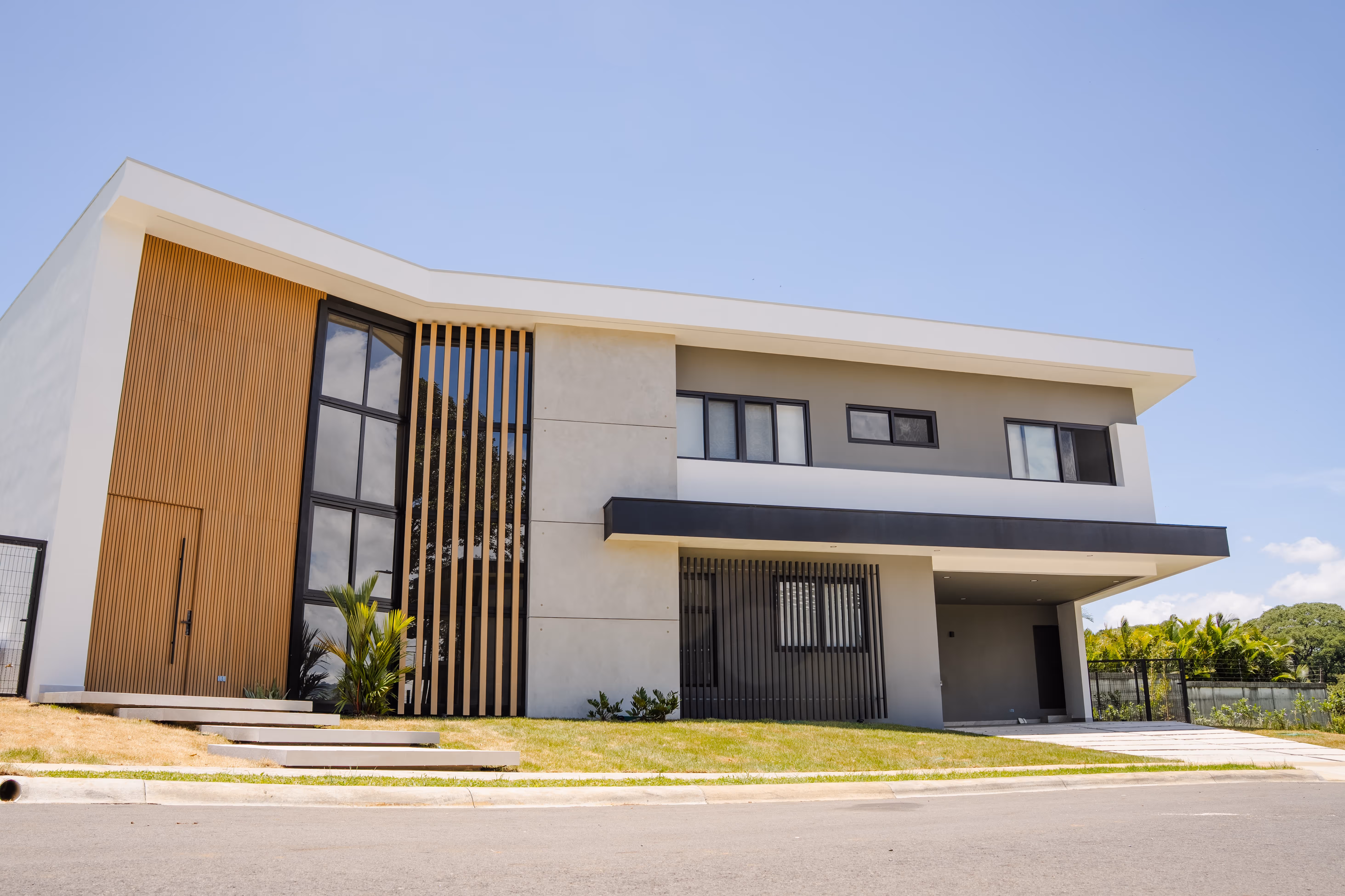 Casa moderna de dos pisos con fachada gris, ventanas negras de pvc, blanca y detalles en madera, césped y cielo azul despejado.