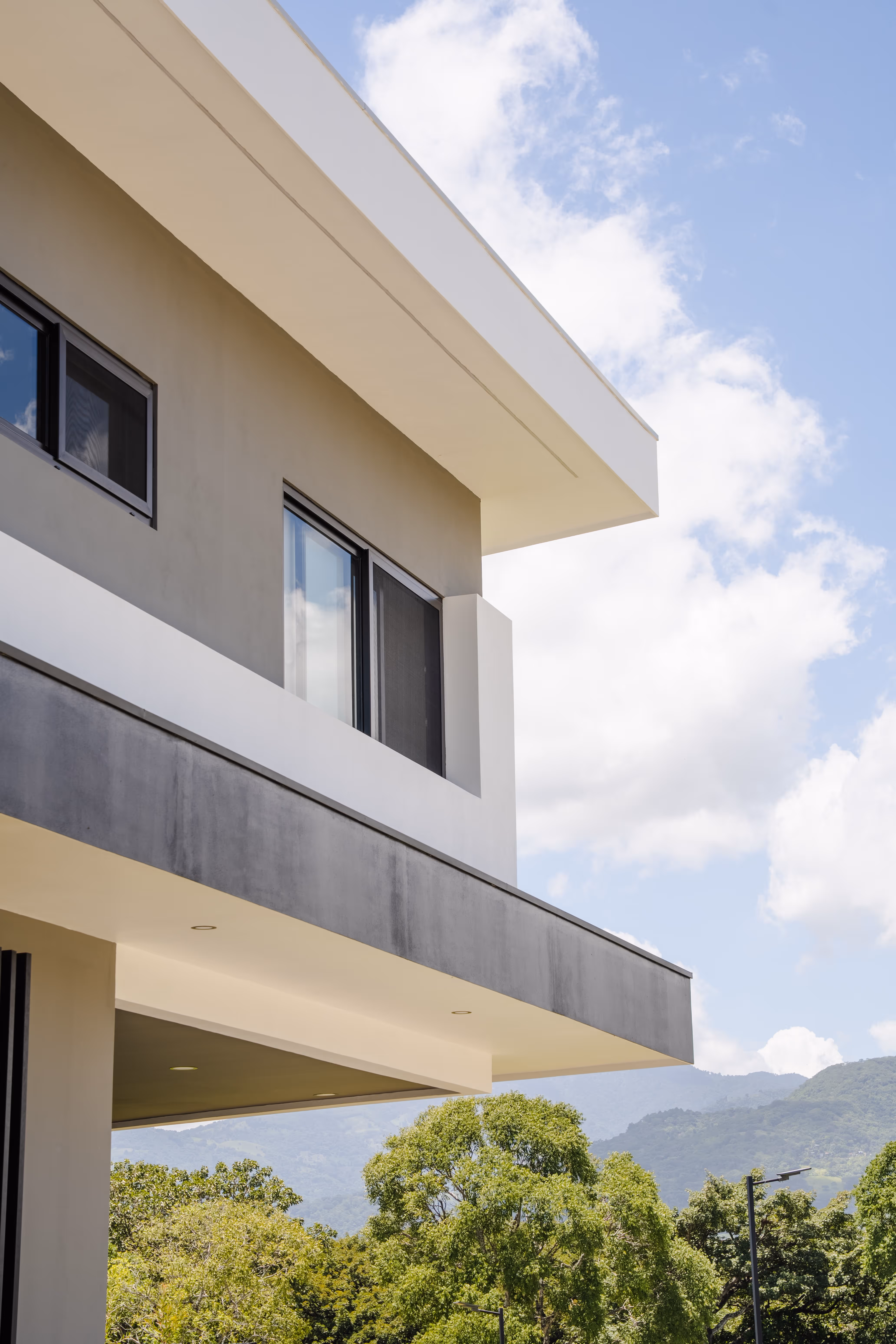 Vista de un edificio moderno con ventanas grandes de PVC negro, árboles verdes y montañas al fondo bajo un cielo parcialmente nublado.