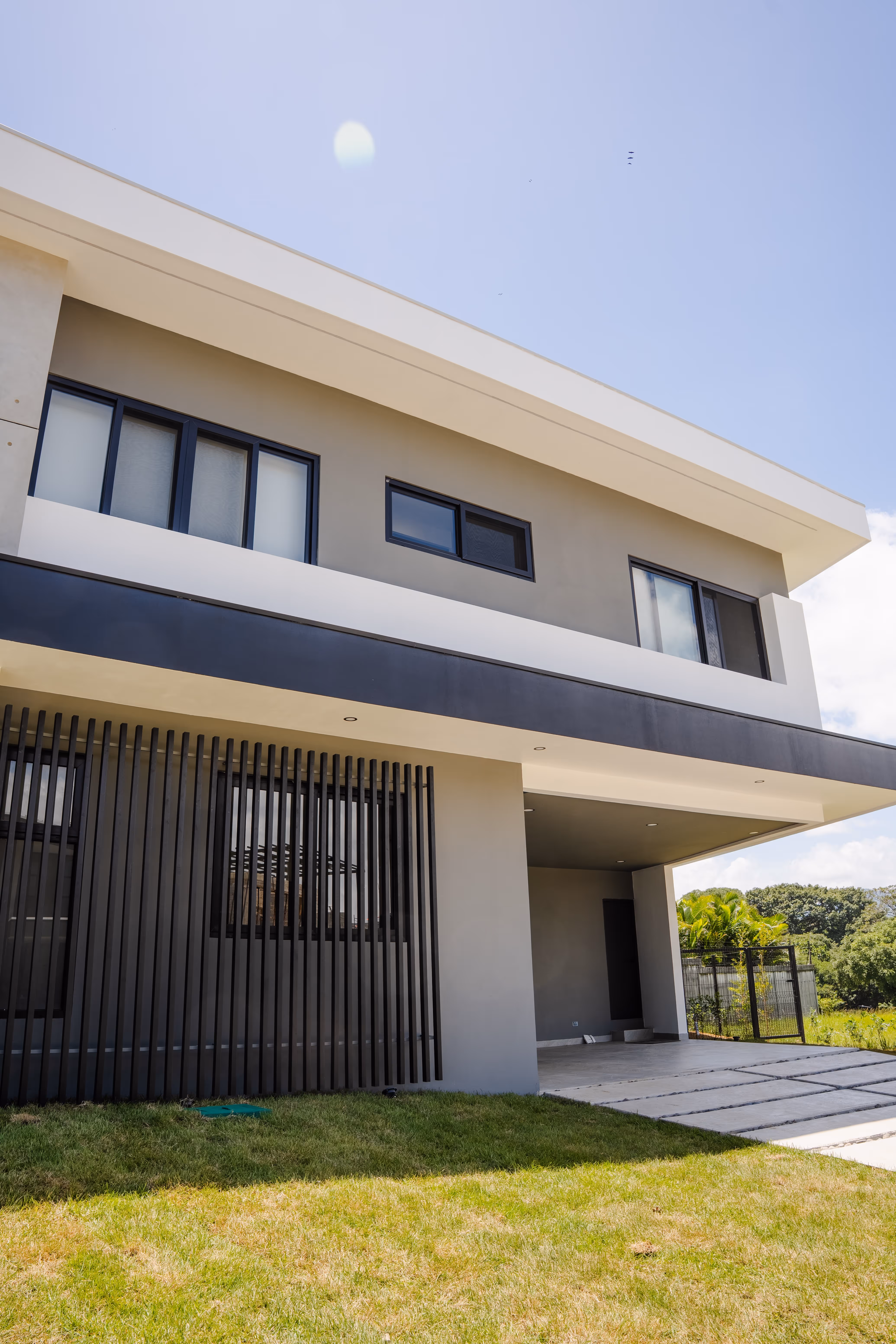 Fachada moderna de dos pisos con ventanas negras de pvc, entrada cubierta y césped verde al frente.