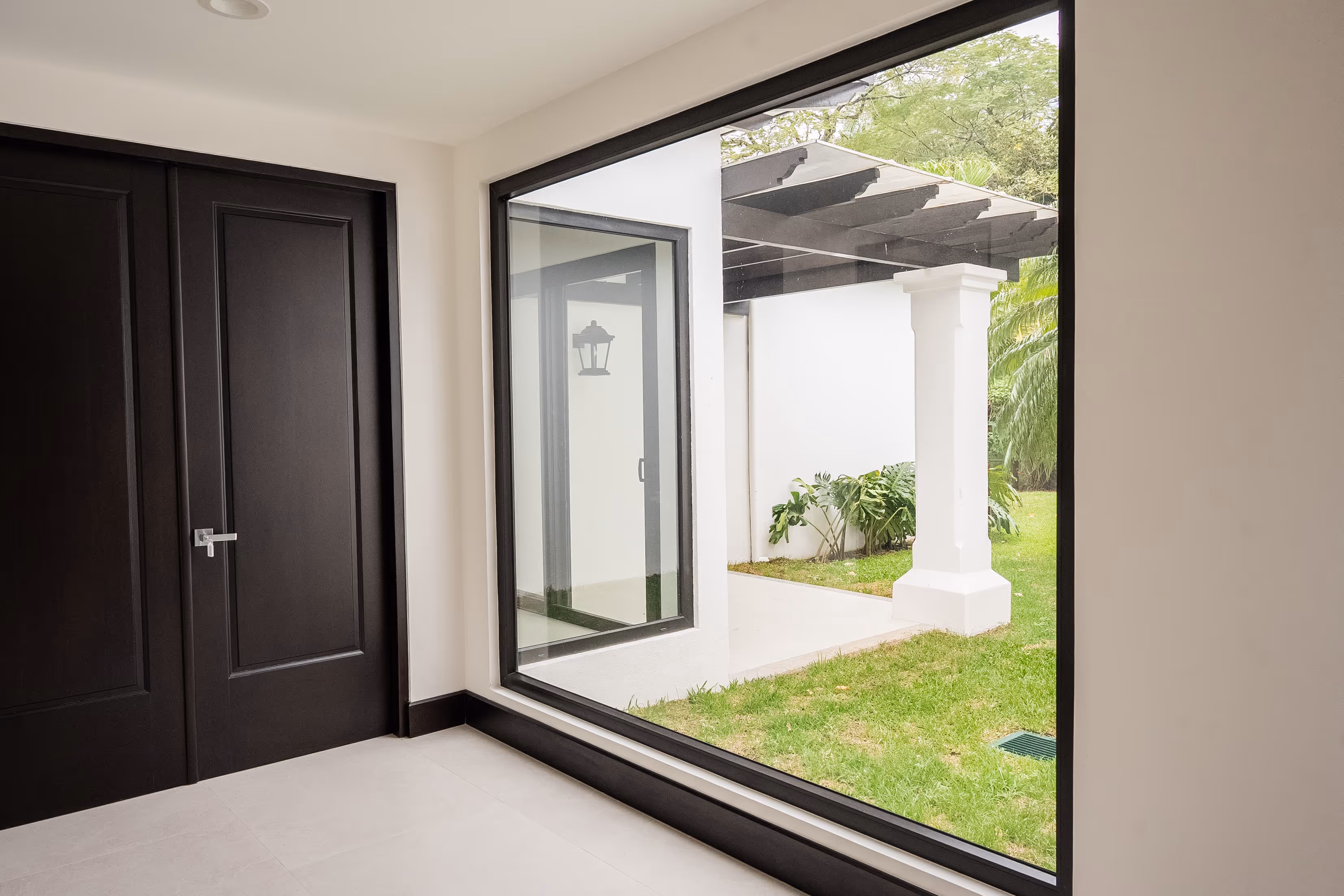 Espacio interior con puertas negras de PVC y gran ventana que muestra un porche con columna blanca y jardín verde.