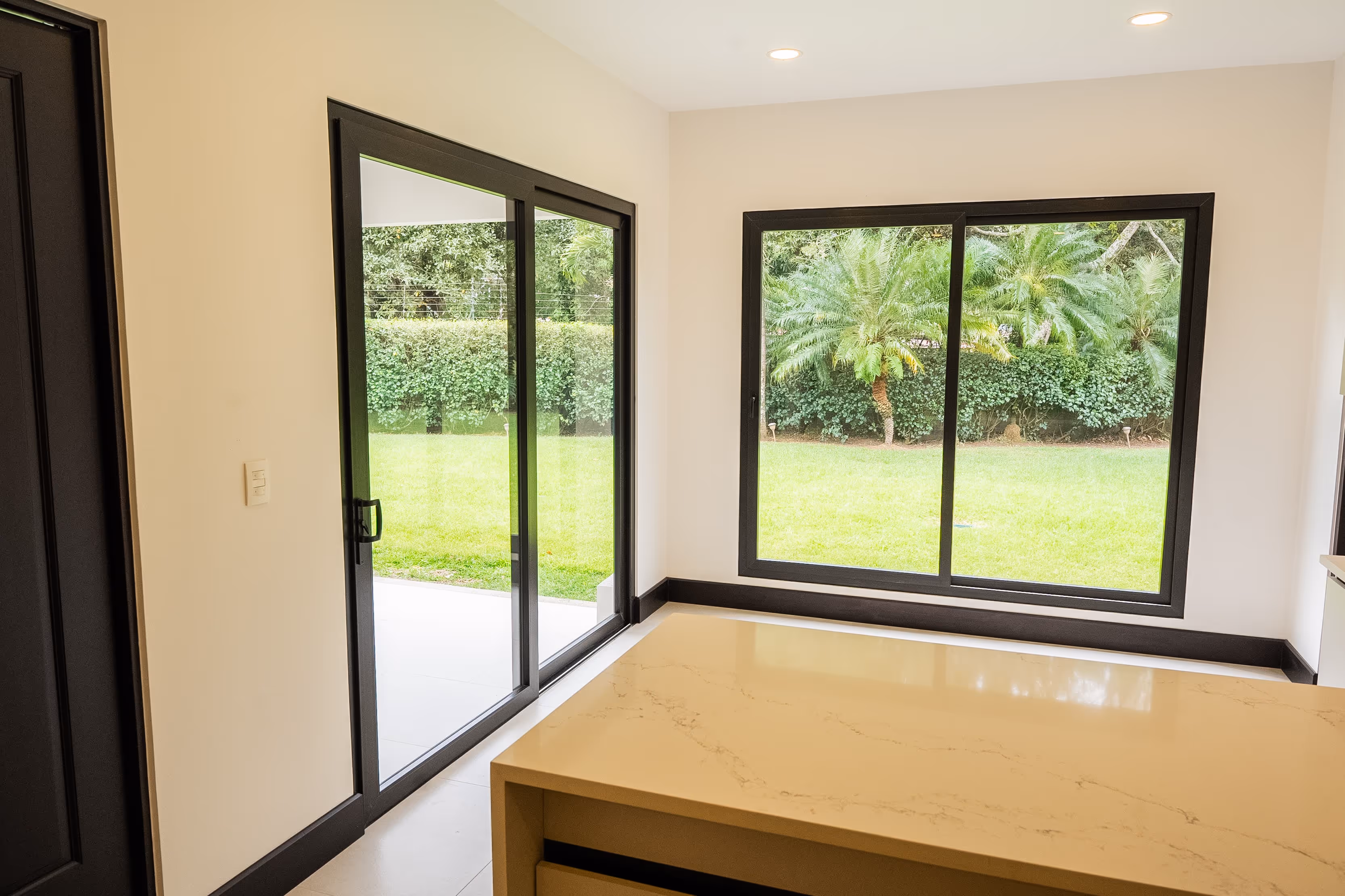 Interior moderno con encimera beige frente a ventanas corredizas de PVC negro que muestran un jardín con césped, setos y palmeras.