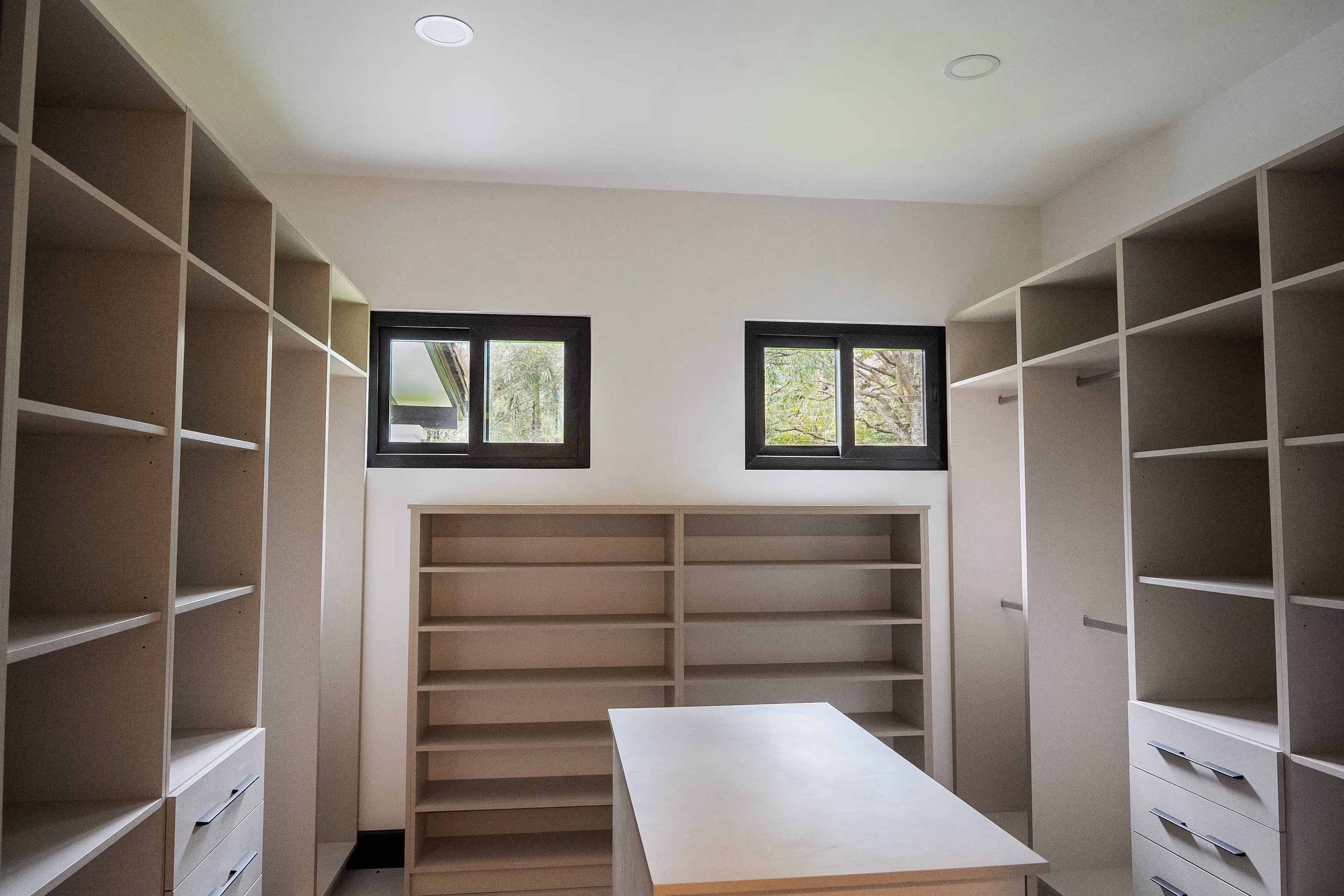 Vestidor amplio con estanterías y cajones blancos, dos ventanas con marco negro de pvc y una mesa central.