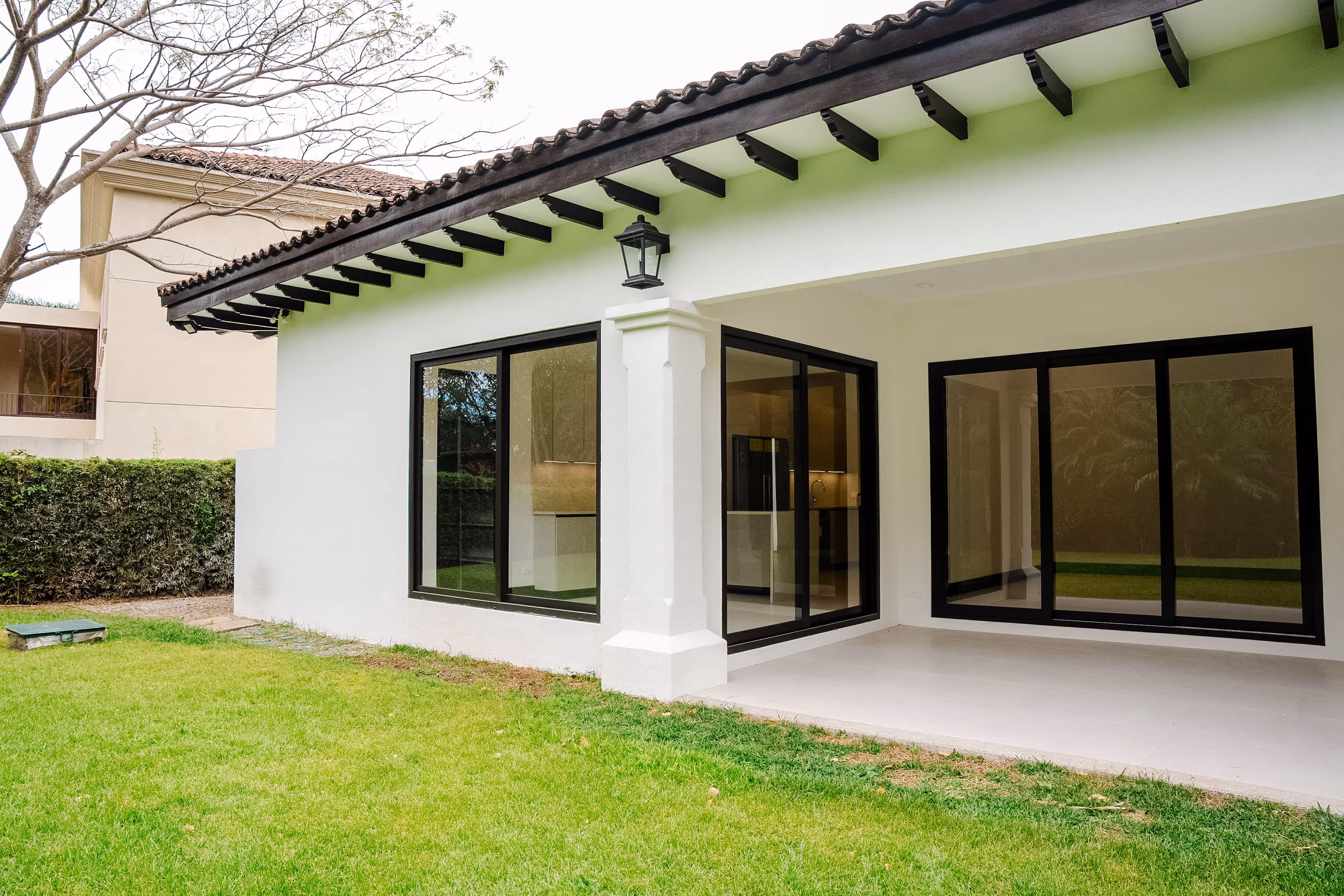 Patio con césped verde y parte trasera de una casa blanca con ventanas corredizas de vidrio y marco negro de PVC.