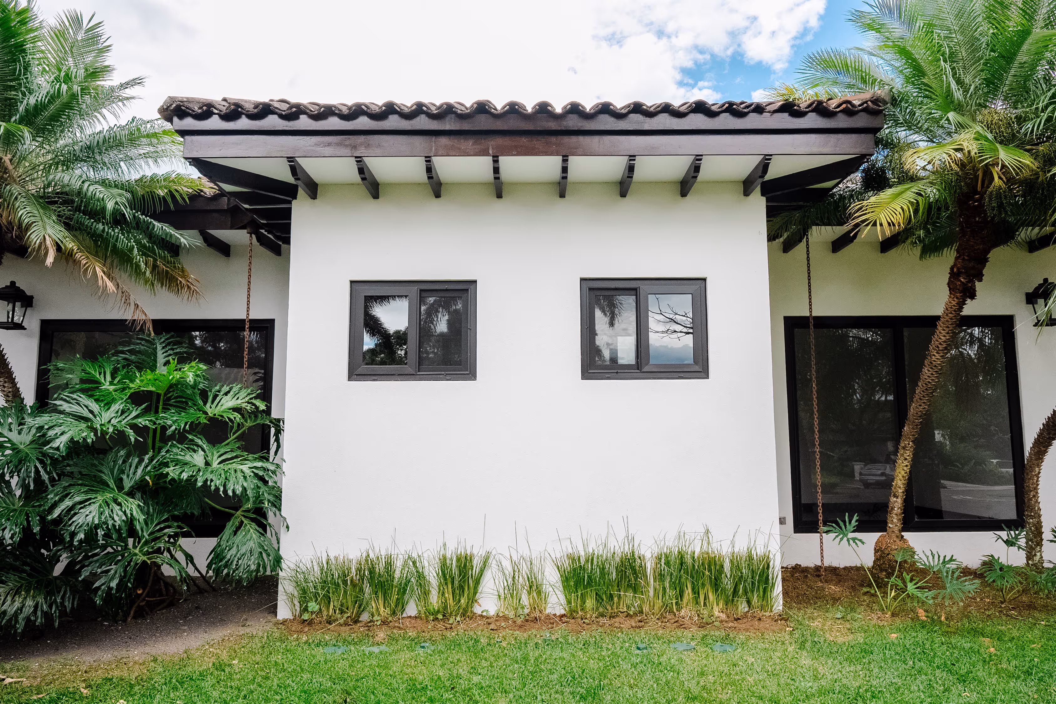 Fachada blanca de una casa con dos ventanas rectangulares de PVC negro, techo de tejas y palmeras alrededor.