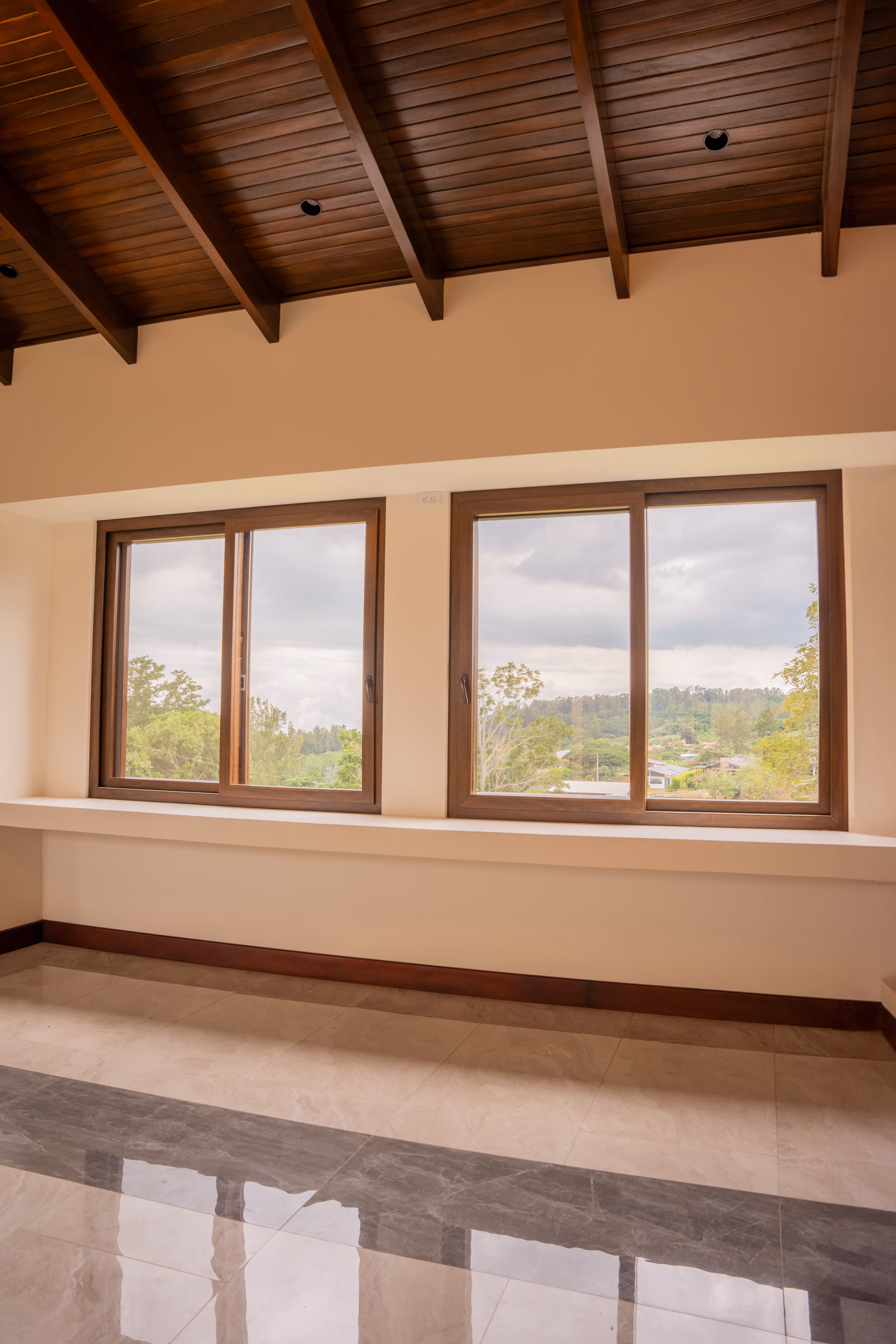 Interior de habitación vacía con techo de madera, suelo de baldosas y ventanas grandes PVC imitación madera con vistas a árboles y cielo nublado.