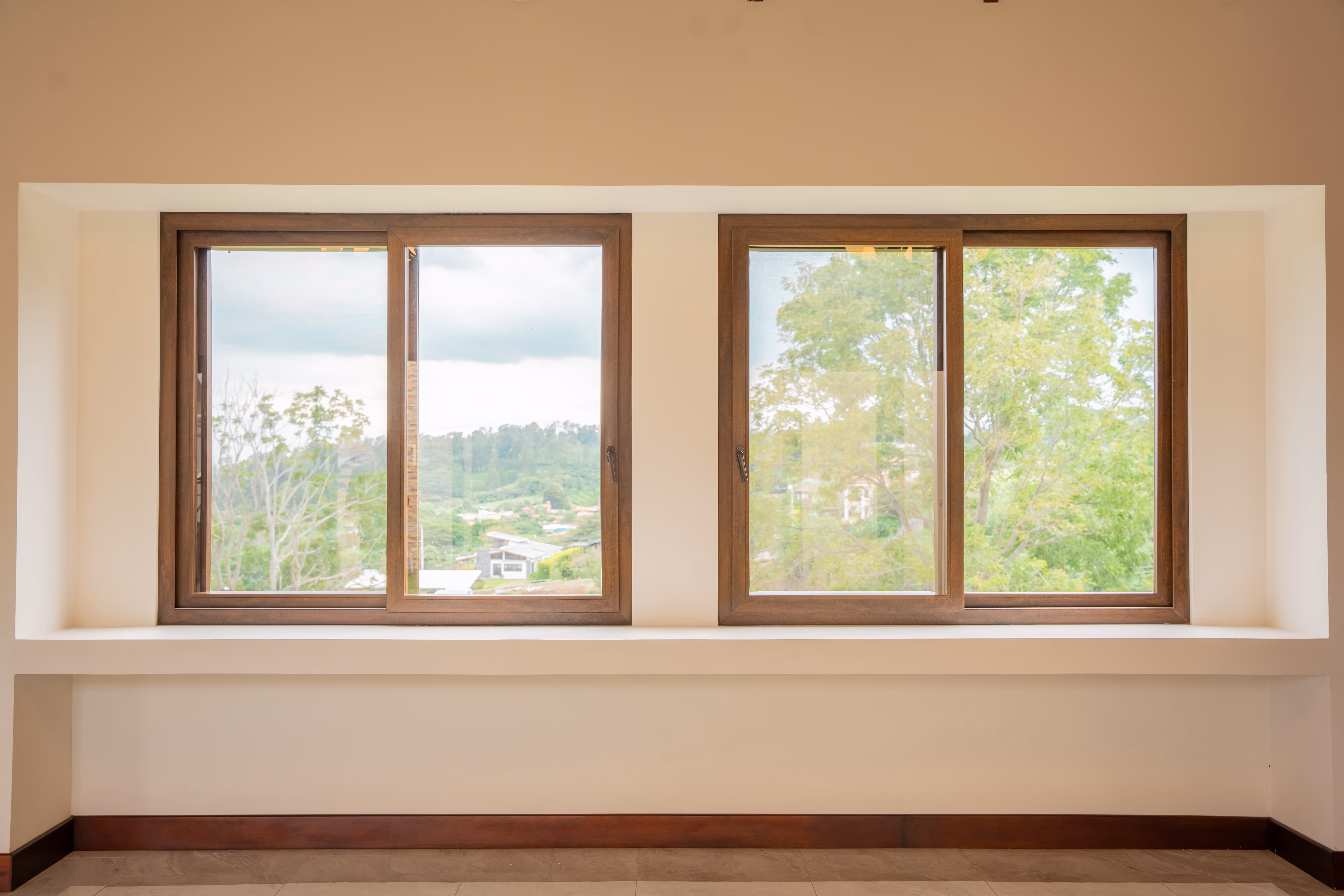 Dos ventanas corredizas de PVC imitación madera en una pared beige con vista a árboles y casas en un paisaje verde.