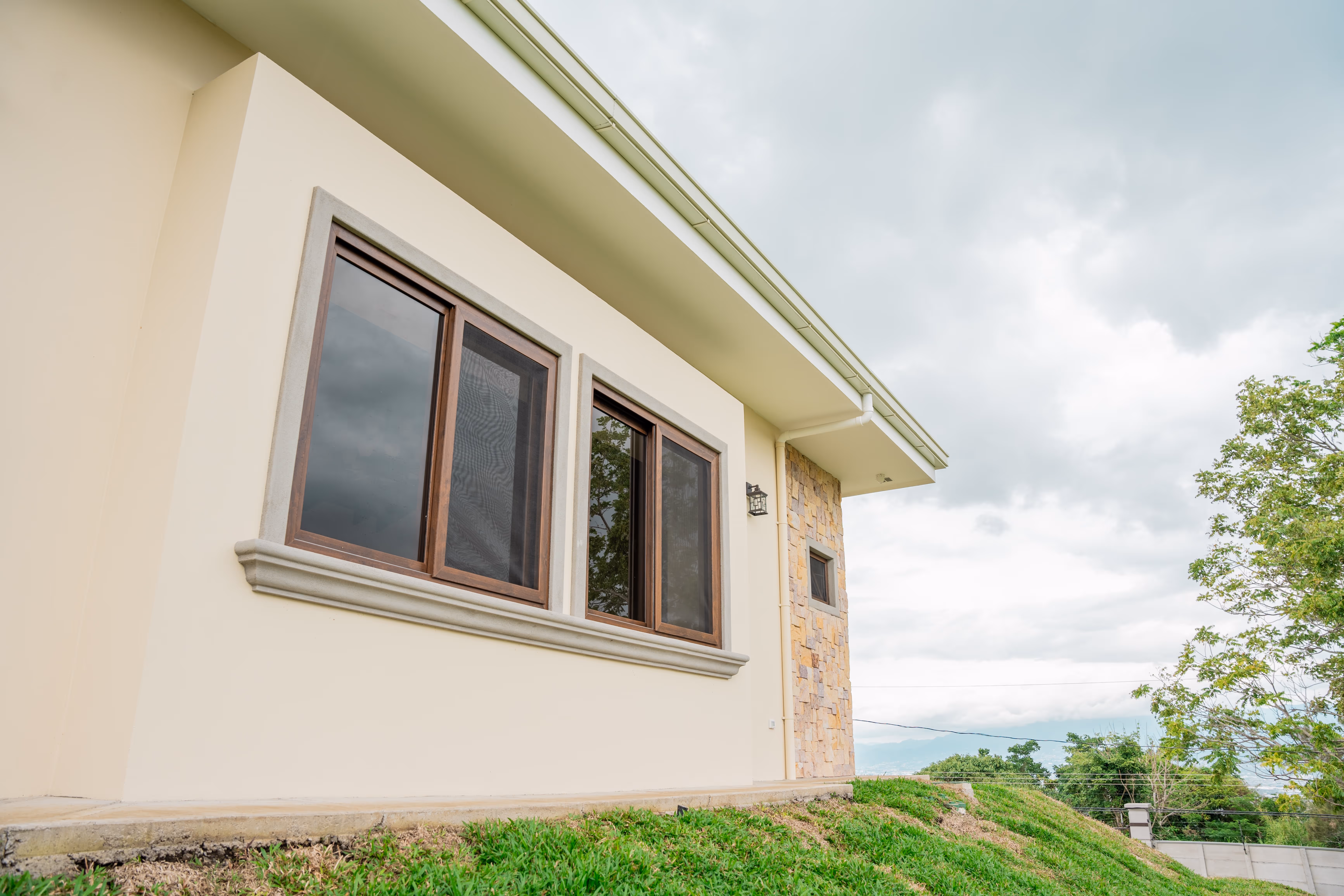 Fachada lateral de casa moderna con ventanas de PVC imitación madera, muro de piedra, césped verde y cielo nublado.