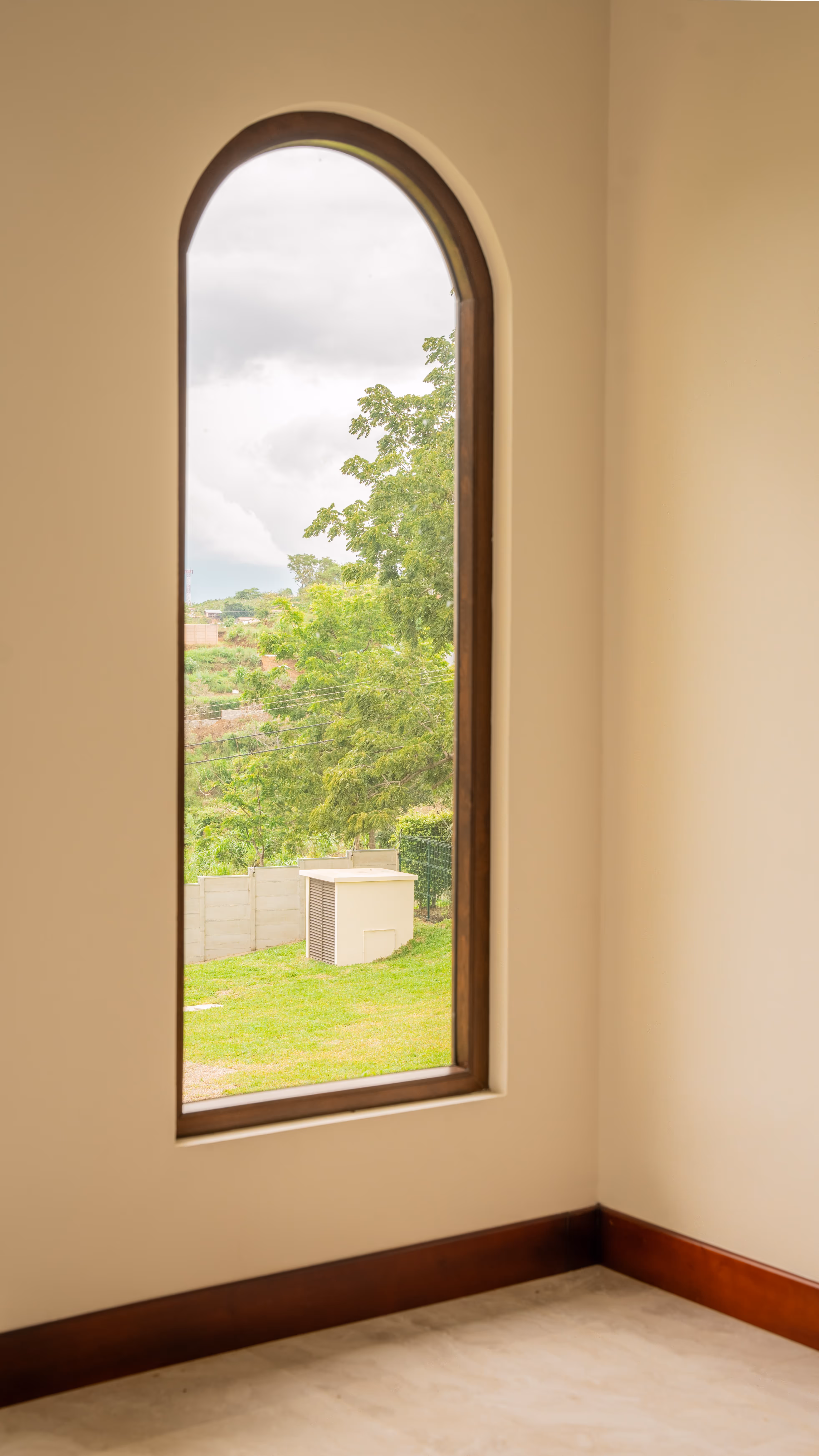 Ventana arqueada con marco de PVC imitación madera que da a un jardín con césped y árboles verdes.