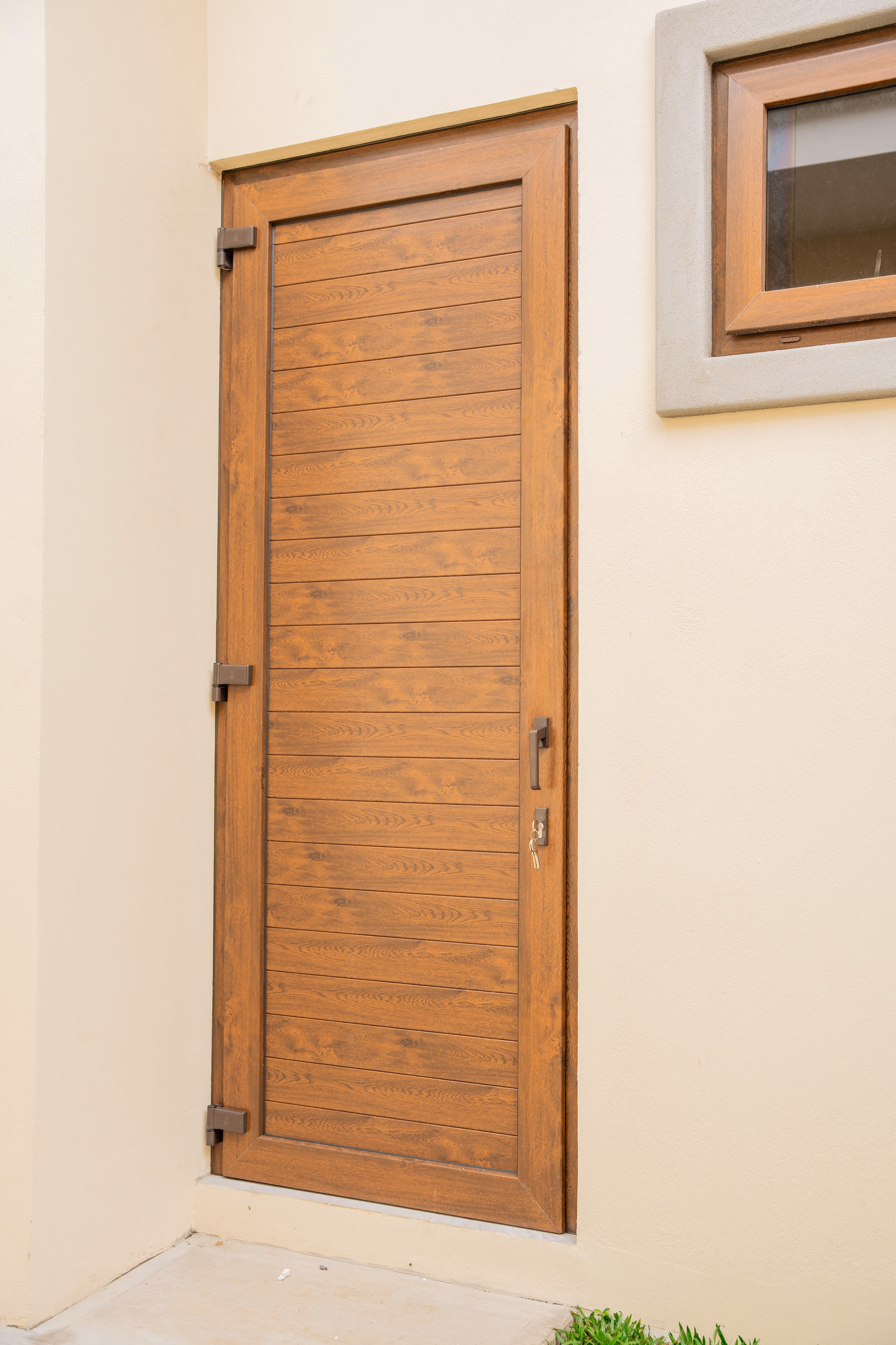 Puerta de PVC imitación madera con marco de madera en pared beige junto a una ventana cuadrada con marco de madera.