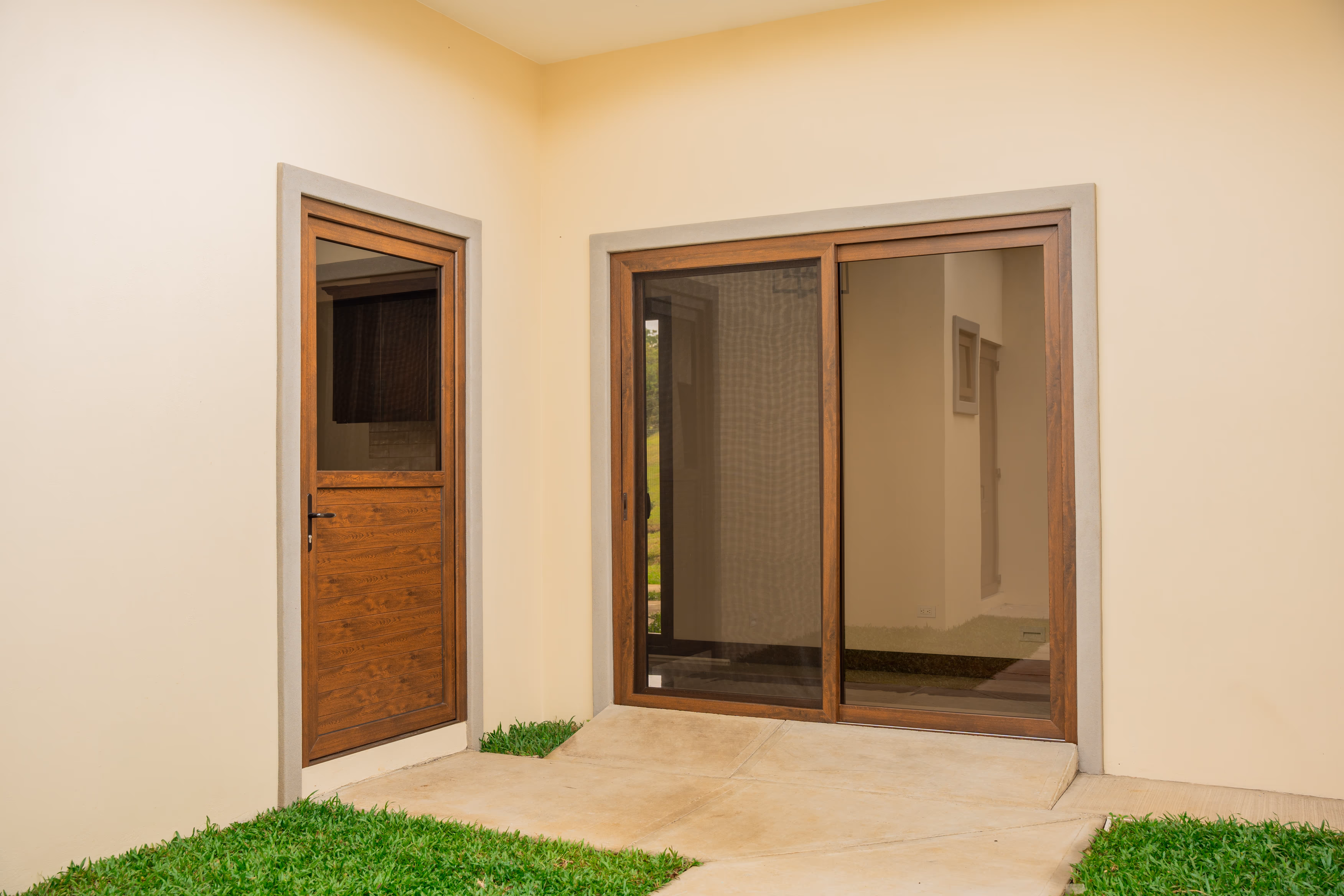 Entrada exterior con puerta de PVC imitación madera y puerta corrediza de vidrio con marco de madera en una pared beige.