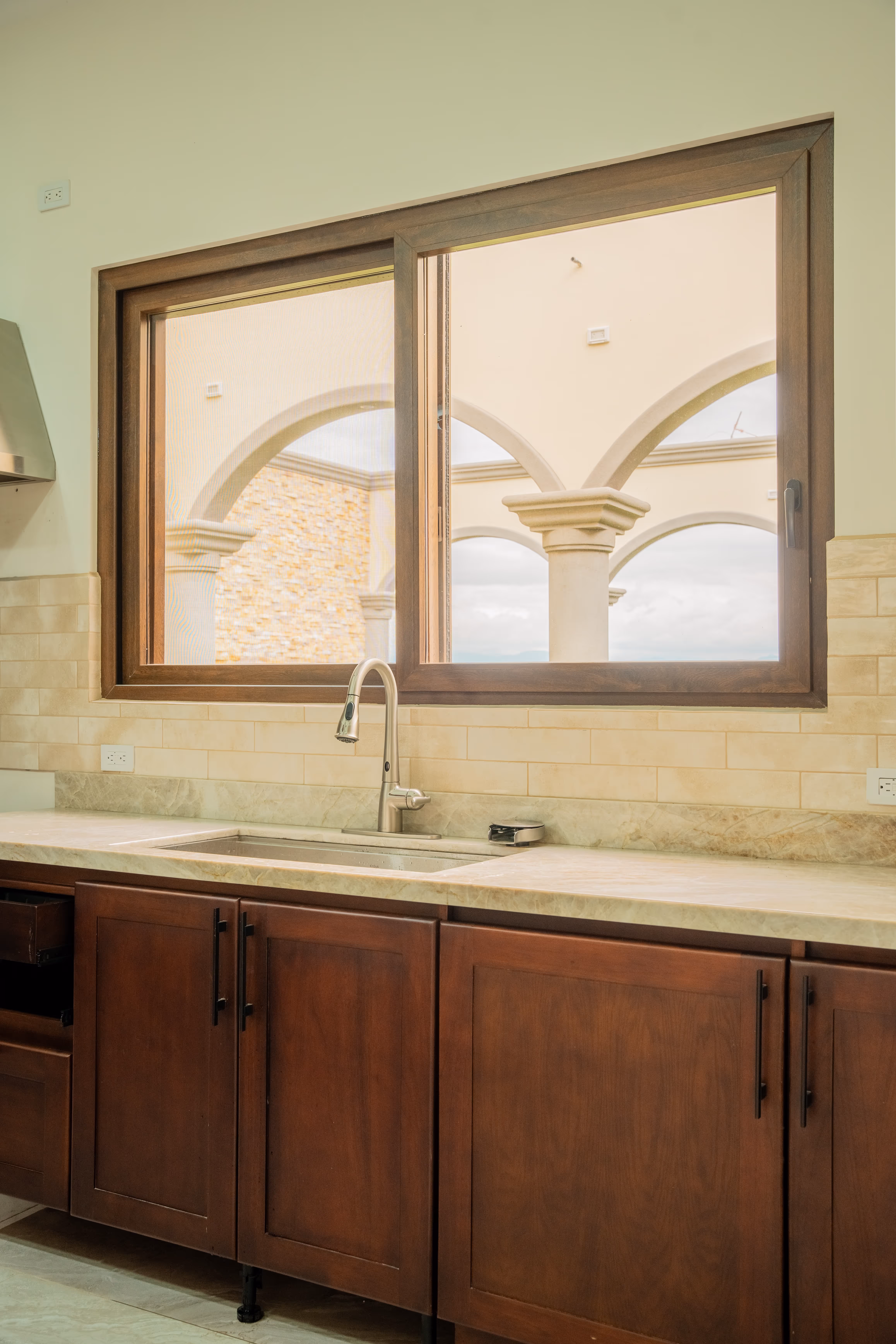 Cocina con encimera de piedra, fregadero de acero inoxidable y armarios de madera oscura bajo una ventana grande de PVC imitación madera con vista a arcos exteriores.