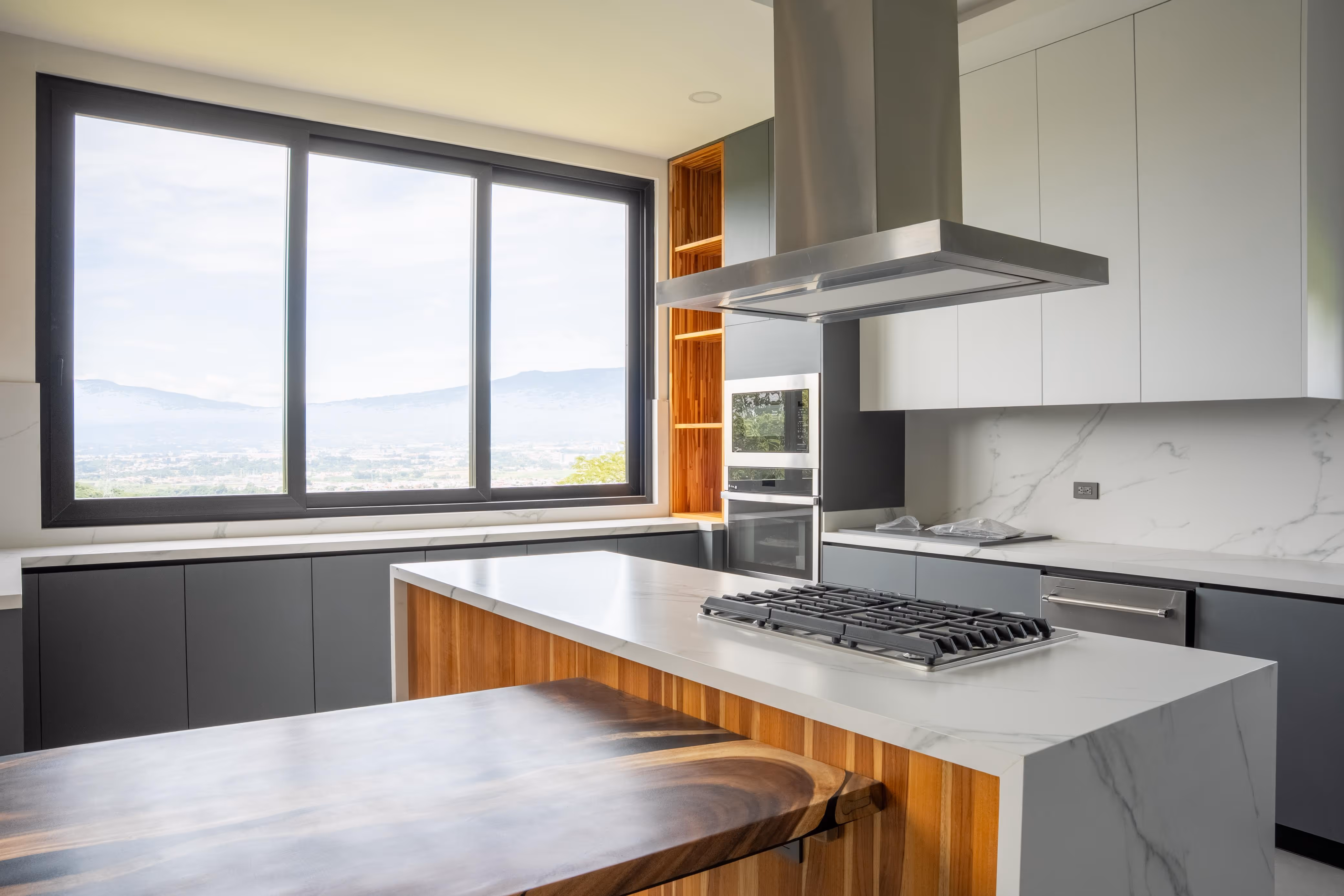 Cocina moderna con encimeras blancas, estufa de gas integrada, campana extractora metálica y una ventana grande color negro de PVC con vista a montañas.