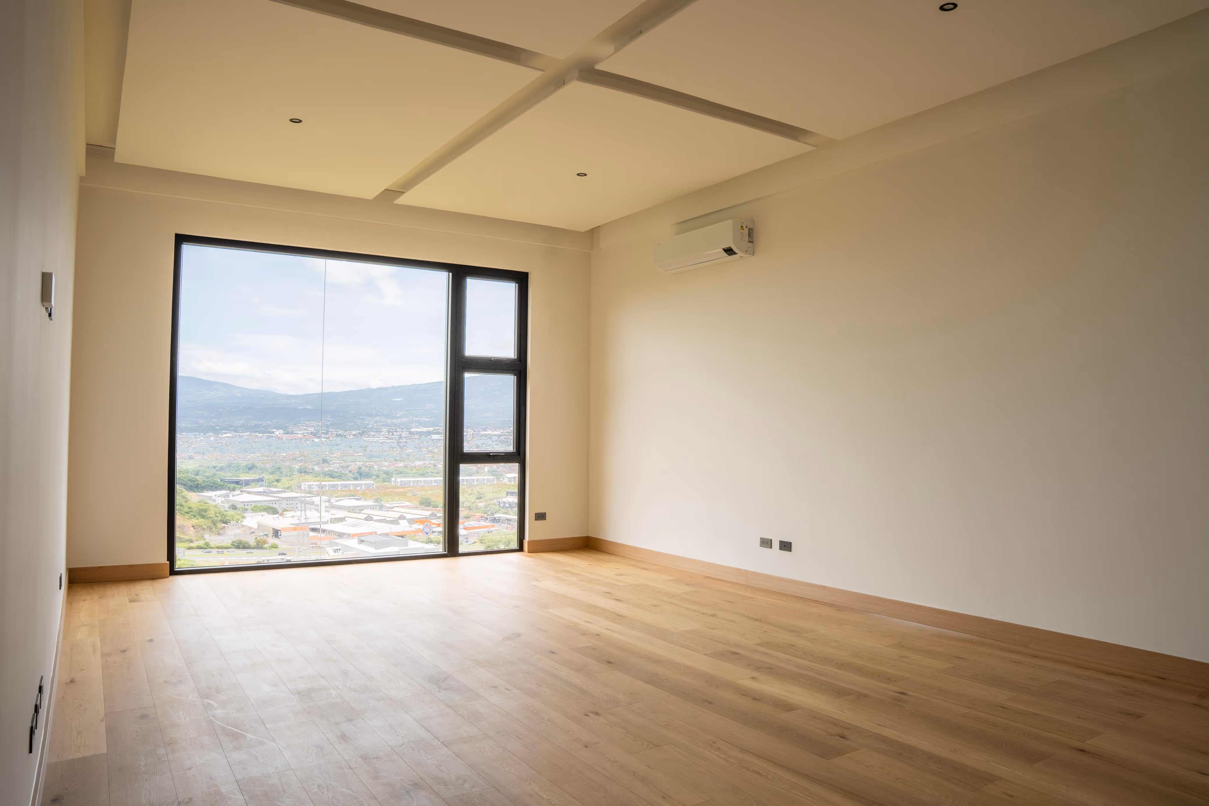 Habitación vacía con piso de madera, ventana grande de PVC con vista a la ciudad y aire acondicionado en la pared.