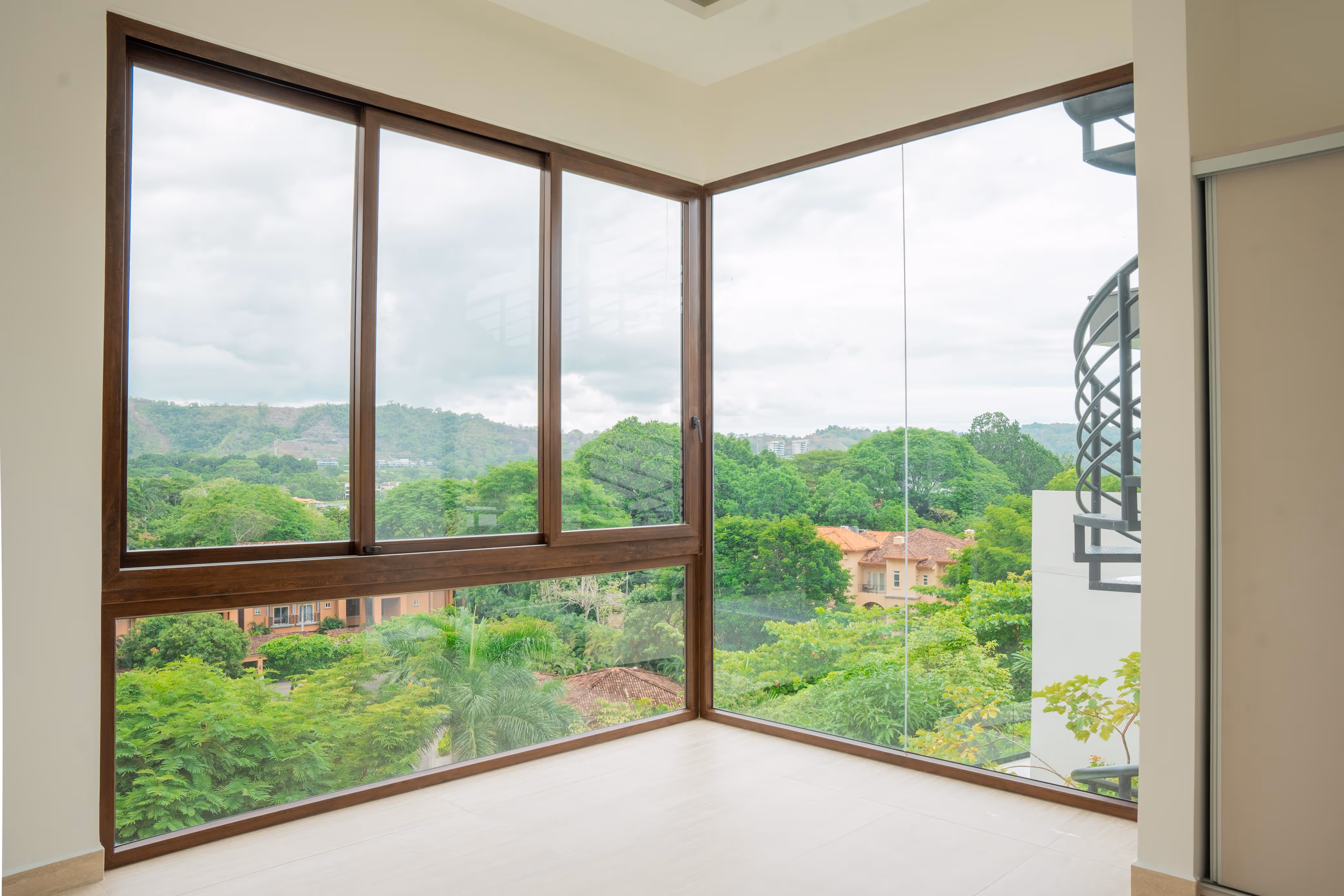 Esquina de habitación con grandes ventanales de PVC imitación madera que muestran una vista de árboles verdes, casas y colinas bajo.