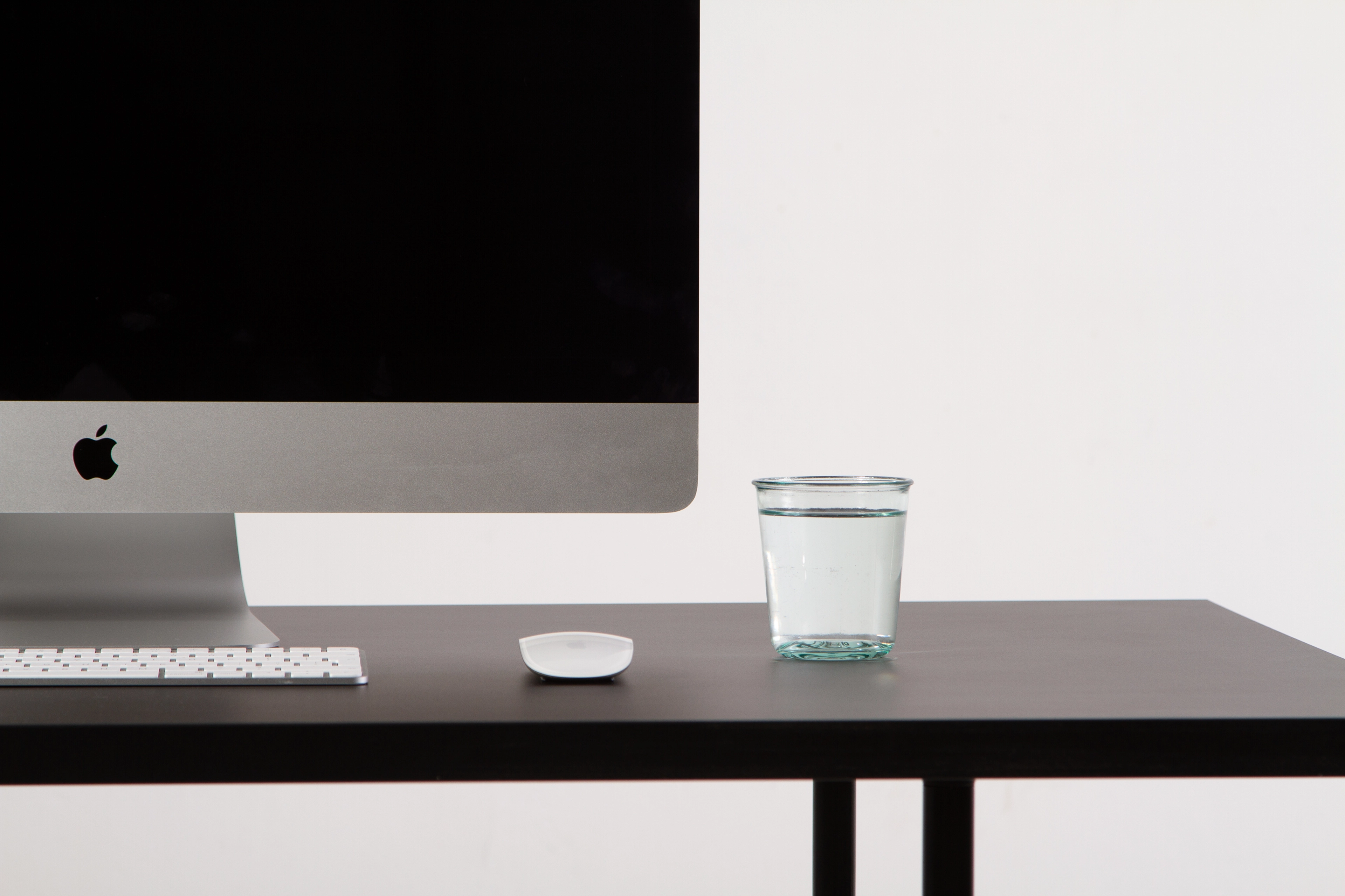 Ciemne biurko z klawiaturą, myszką i szklanką wody obok monitora iMac