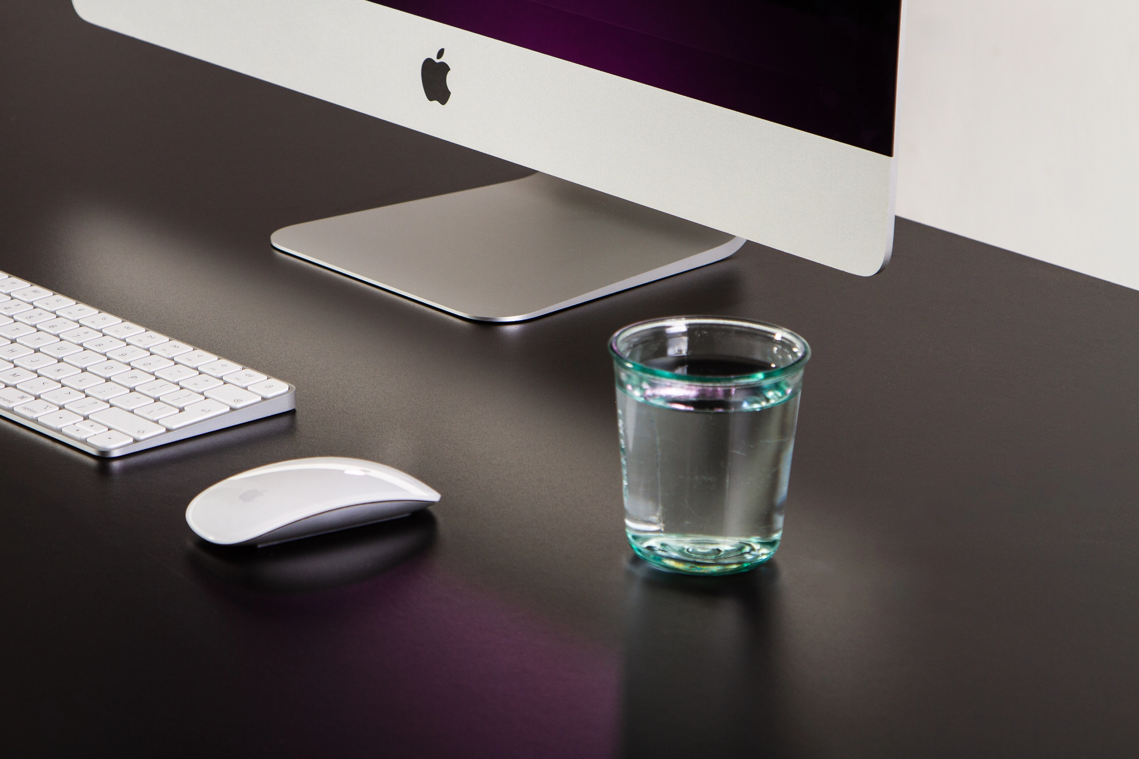 Fragment biurka z klawiaturą, myszką Apple oraz szklanką wody, w tle monitor iMac