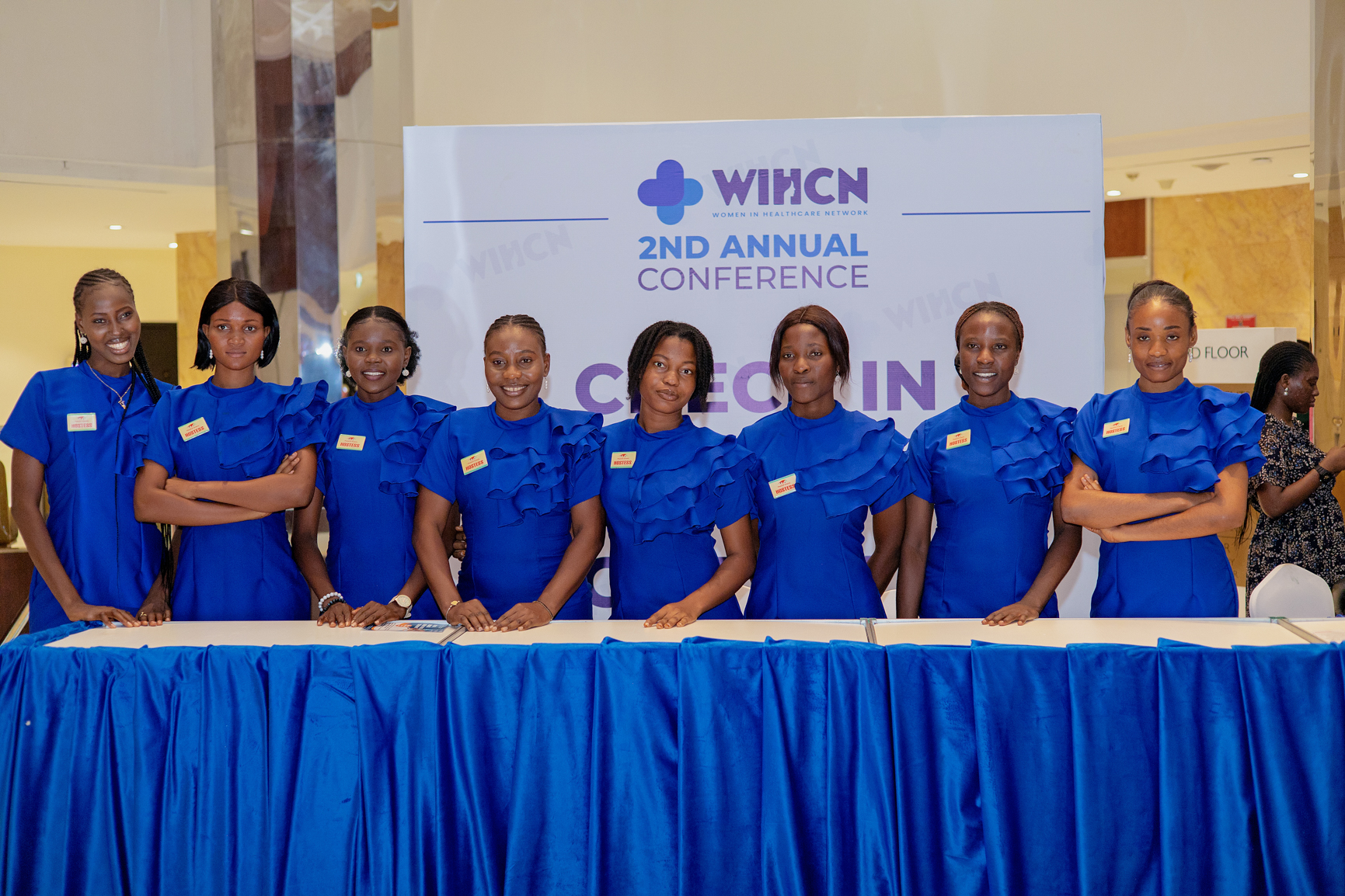 Eight women in matching blue dresses standing behind a blue-draped table at the Women in Healthcare Network 2nd Annual Conference check-in desk.