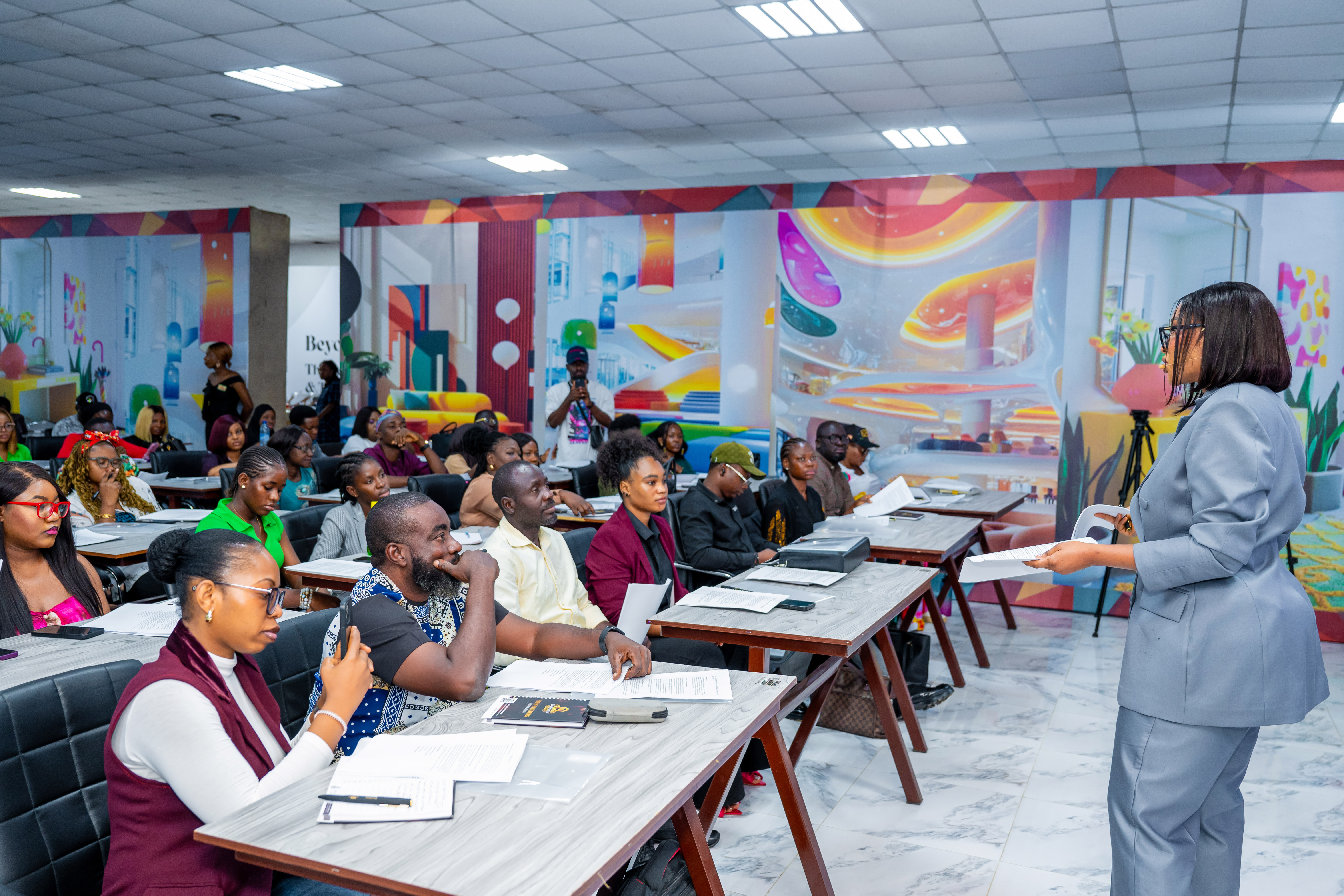 Woman in a gray suit presenting to a diverse group of attentive people seated at desks in a brightly decorated conference room.