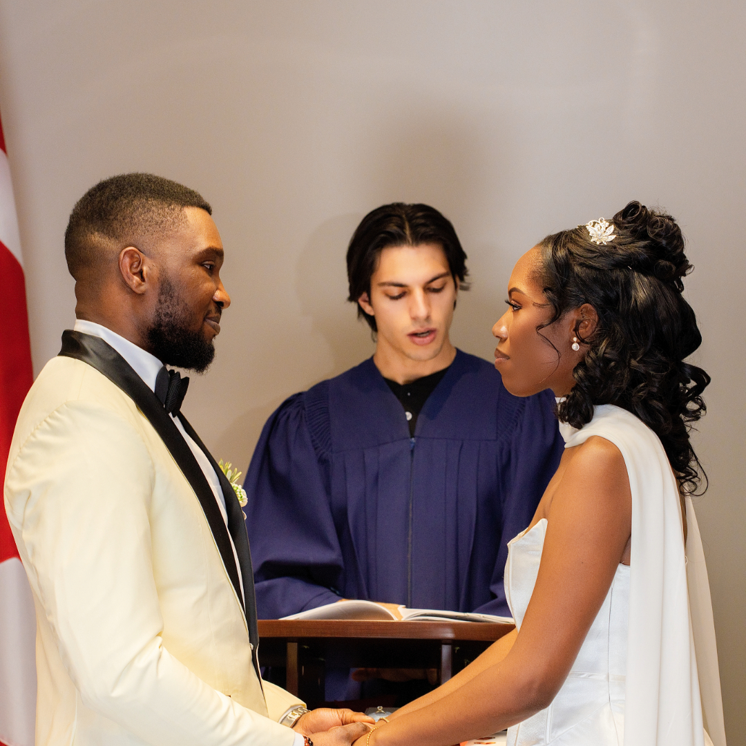 Groom in white tuxedo and bride in white dress holding hands during wedding ceremony officiated by young person in purple robe.