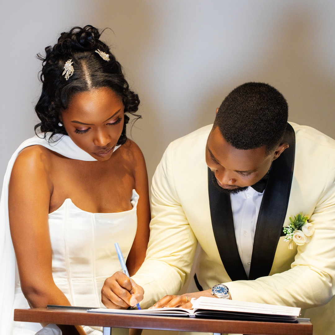 Bride in white dress and groom in light suit signing a document together at a table.