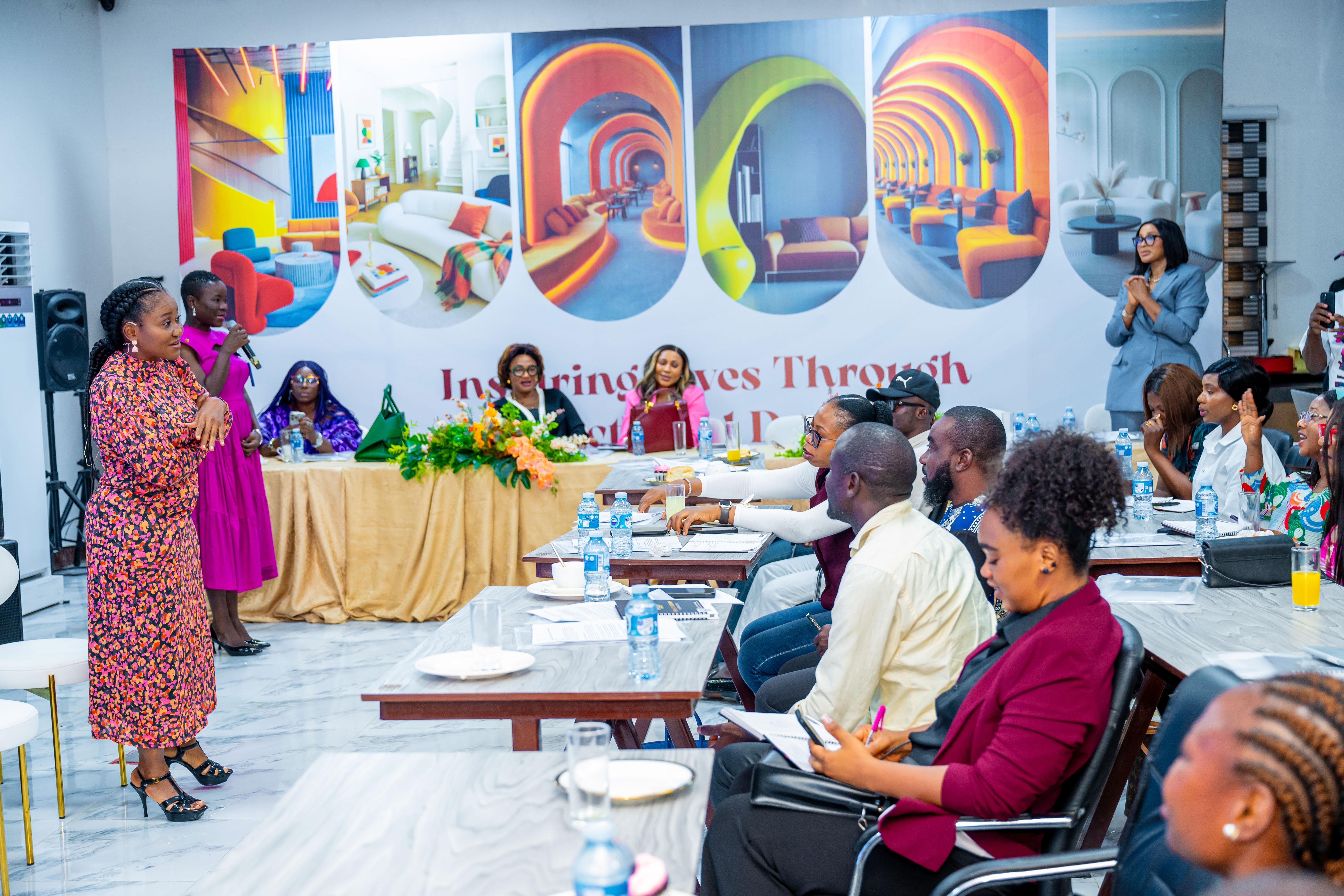 A woman in a floral dress speaking to an audience seated at tables in a brightly lit room with colorful wall decor and several people taking notes or listening.