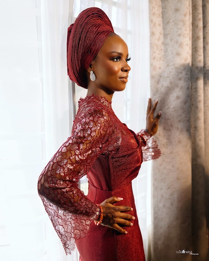 Side profile of a woman dressed in an elegant burgundy lace outfit and matching headwrap, standing by a window with her hand on the curtain.