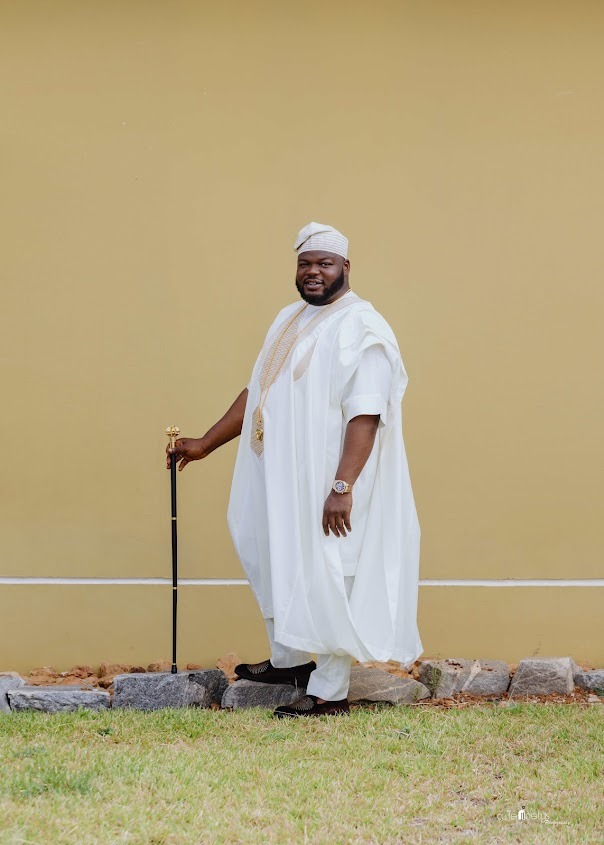 Man dressed in traditional white agbada attire with a matching cap, standing outdoors on grass near rocks, holding a black cane with a gold handle.
