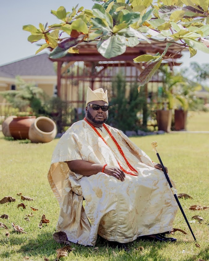 Man wearing traditional cream and gold attire with red beads and sunglasses, sitting on grass holding a black cane with a gold handle.