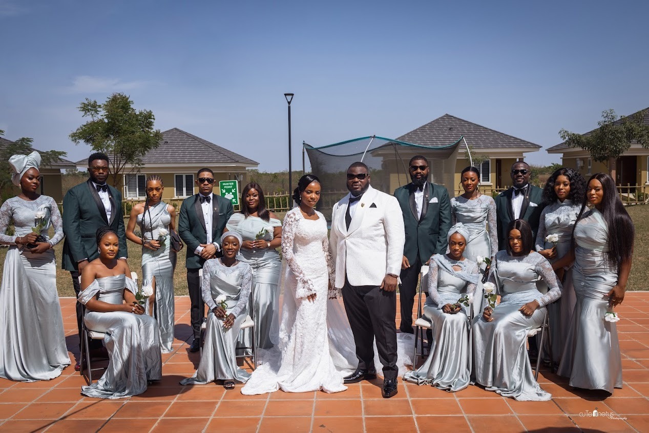Bride in white lace gown and groom in white jacket with groomsmen and bridesmaids in matching silver attire posing outdoors on tiled ground.