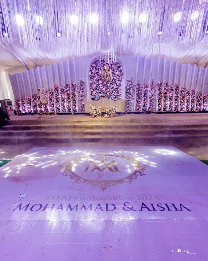 Wedding stage decorated with pink and white flowers, white sofa, and illuminated ceiling drapes; floor displays 'Mohammad & Aisha' and '#MAlroyalwedding2023'.