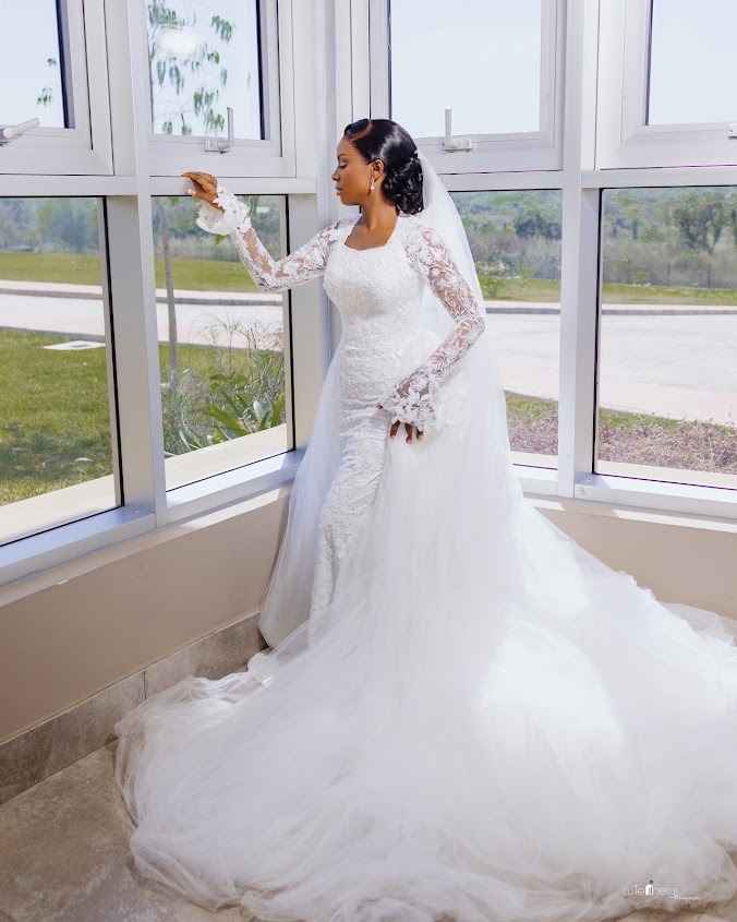 Bride in a white lace and tulle wedding gown with long sleeves standing by a large window looking outside.