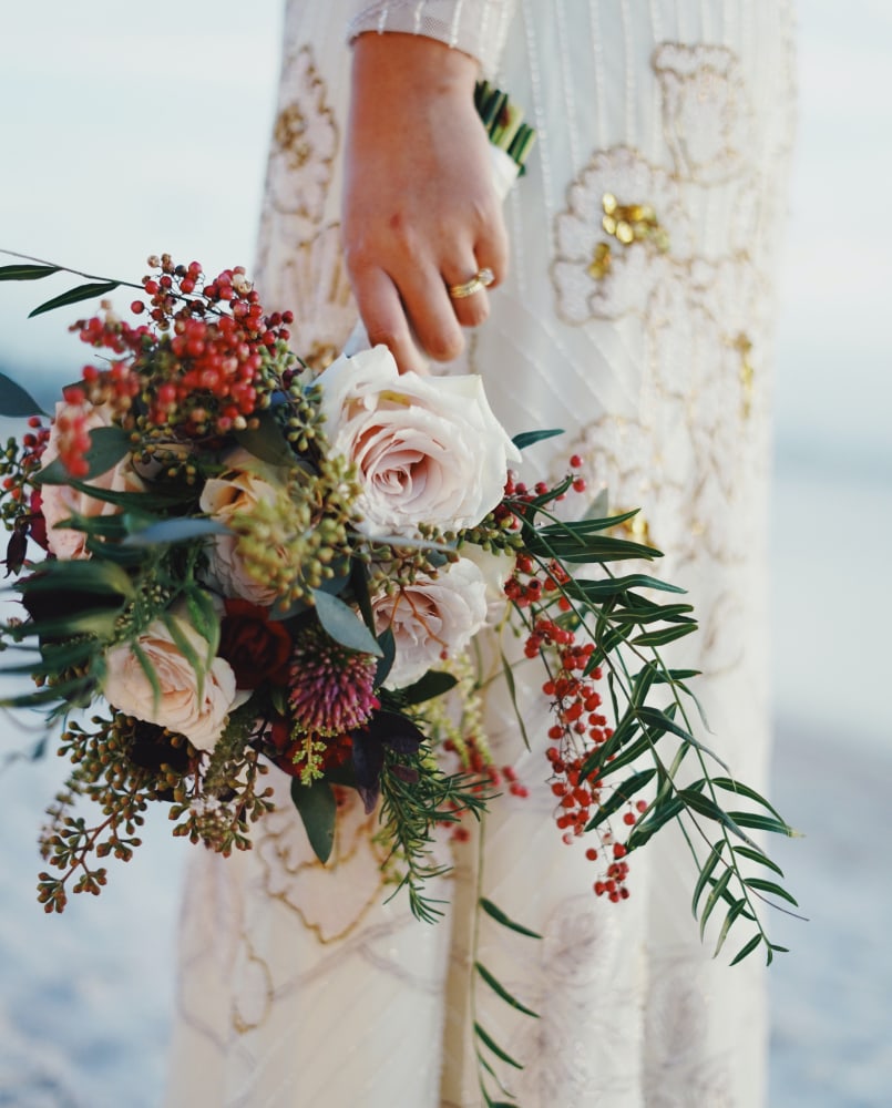 Braut hält einen Blumenstrauss mit weissen Rosen, roten Beeren und grünem Laub, vor einem weiss-gold bestickten Kleid.