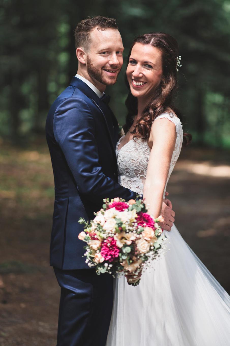 Glückliches Brautpaar lächelt in die Kamera, sie hält einen bunten Blumenstrauß, Nahaufnahme im Wald.