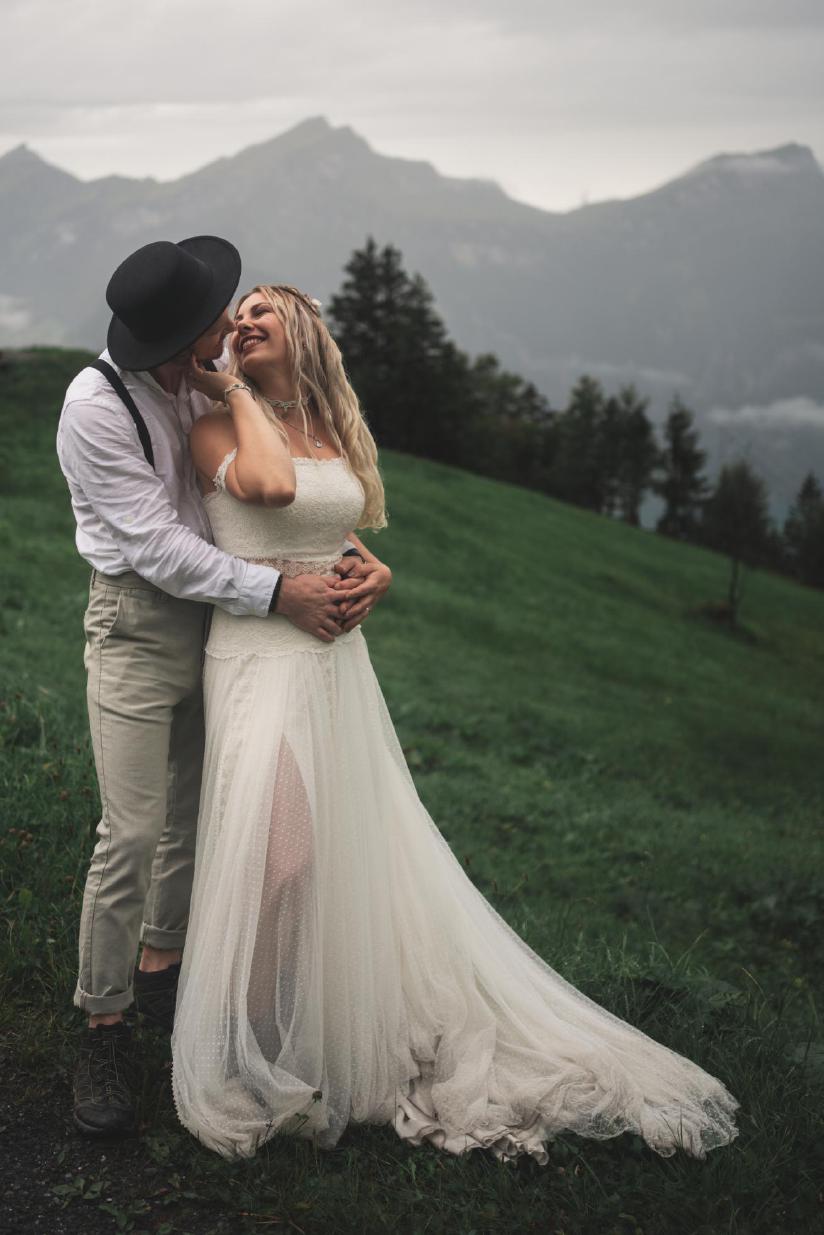 Glückliches Brautpaar in Ganzkörperaufnahme auf einer grünen Bergwiese unter bewölktem Himmel.