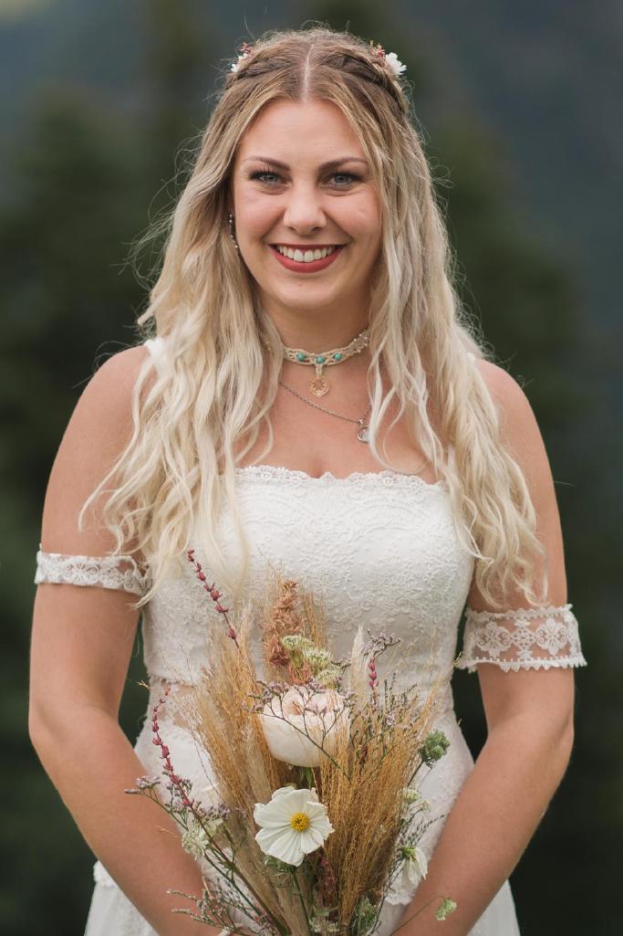 Porträt einer lächelnden Boho-Braut mit langen blonden Locken, die ein weißes, trägerloses Hochzeitskleid mit Spitze und einen natürlichen, getrockneten Blumenstrauß in den Händen hält. Im Hintergrund ist eine unscharfe, grüne Outdoor-Landschaft zu sehen.